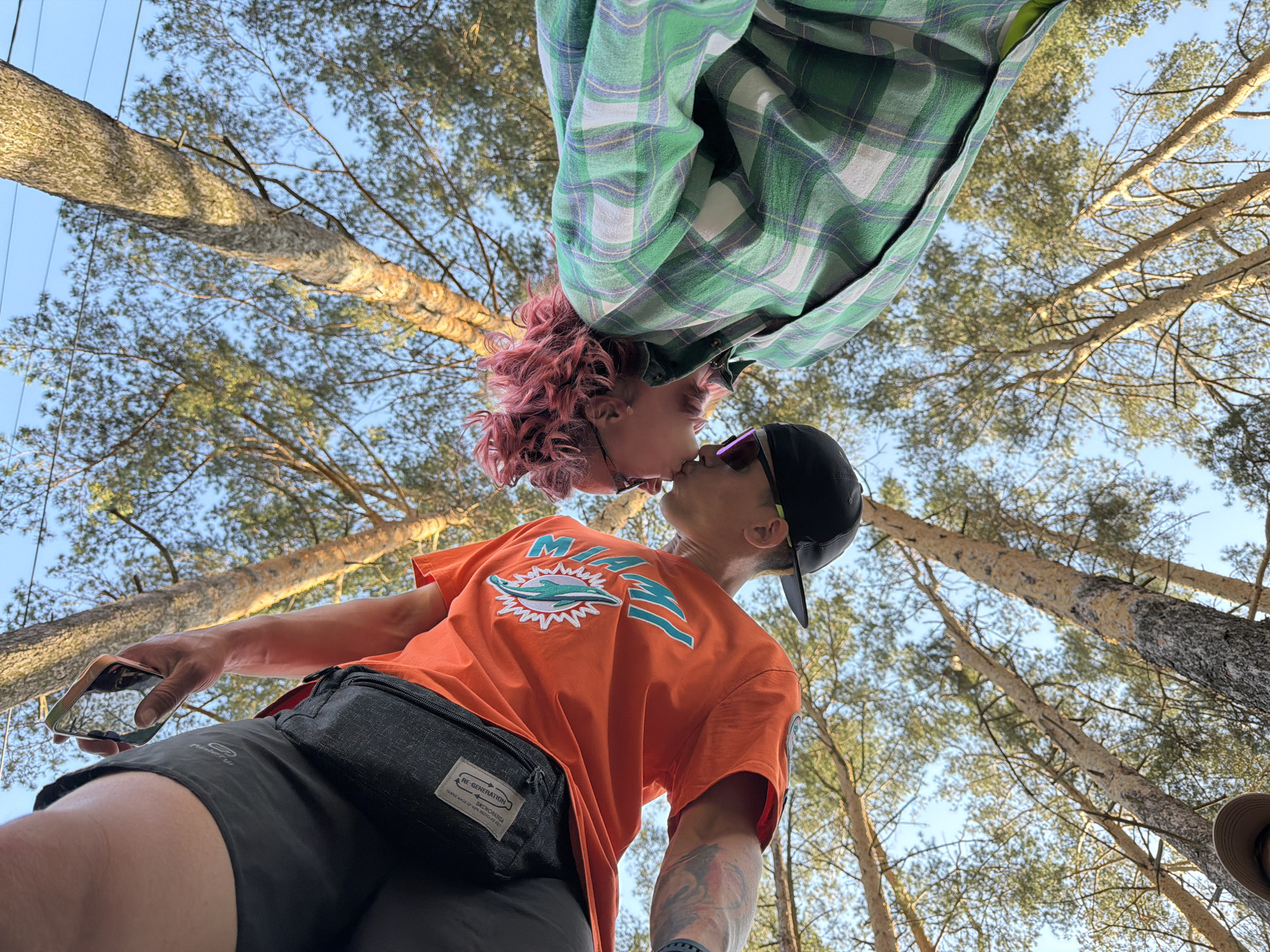 From a low angle, a couple is seen kissing outdoors. One person has pink curly hair and is wearing a green and white plaid shirt. The other person wears an orange t-shirt, black shorts with a black waist bag, a black baseball cap, and sunglasses. Tattoos are visible on the arm of the person in the orange shirt, and they are holding a cell phone in one hand. Above them, trees reach towards a blue sky. Based on the image metadata, the location is Chaikovsky, Russia.