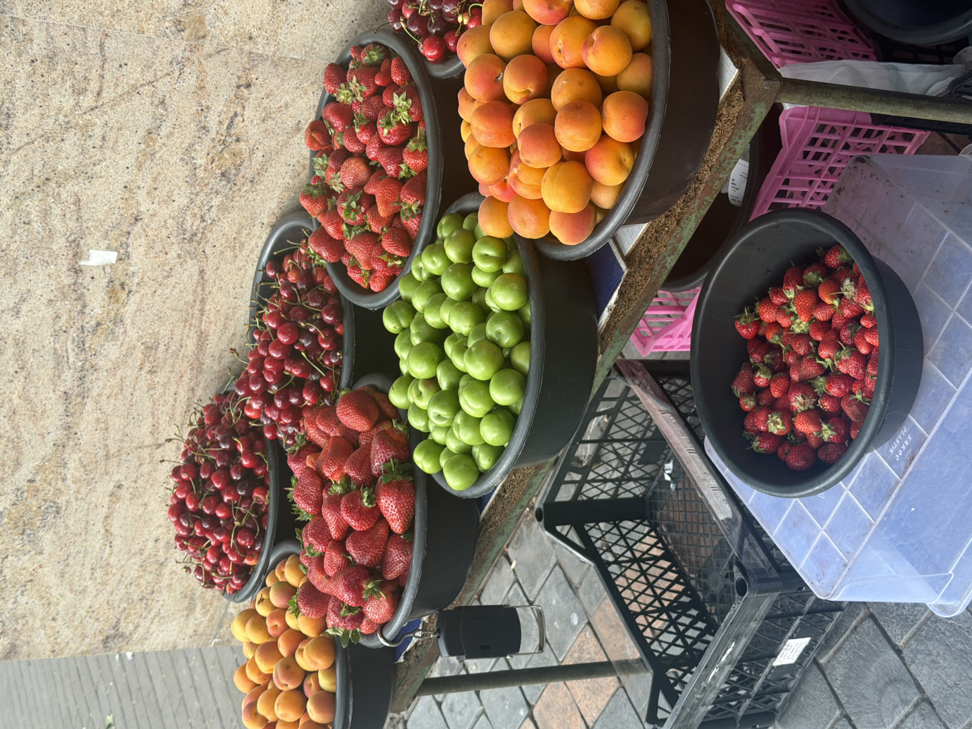 The image shows a fruit stand on a street in Yerevan, Armenia. The stand is stocked with a variety of fresh fruits, including strawberries, cherries, apricots, and green plums, all displayed in black plastic containers. The fruits appear vibrant and ripe. The stand itself is constructed of metal and has a tiered design, with plastic crates stacked beneath the fruit containers. The background includes a textured sidewalk and glimpses of the urban environment. No people are visible in the image.