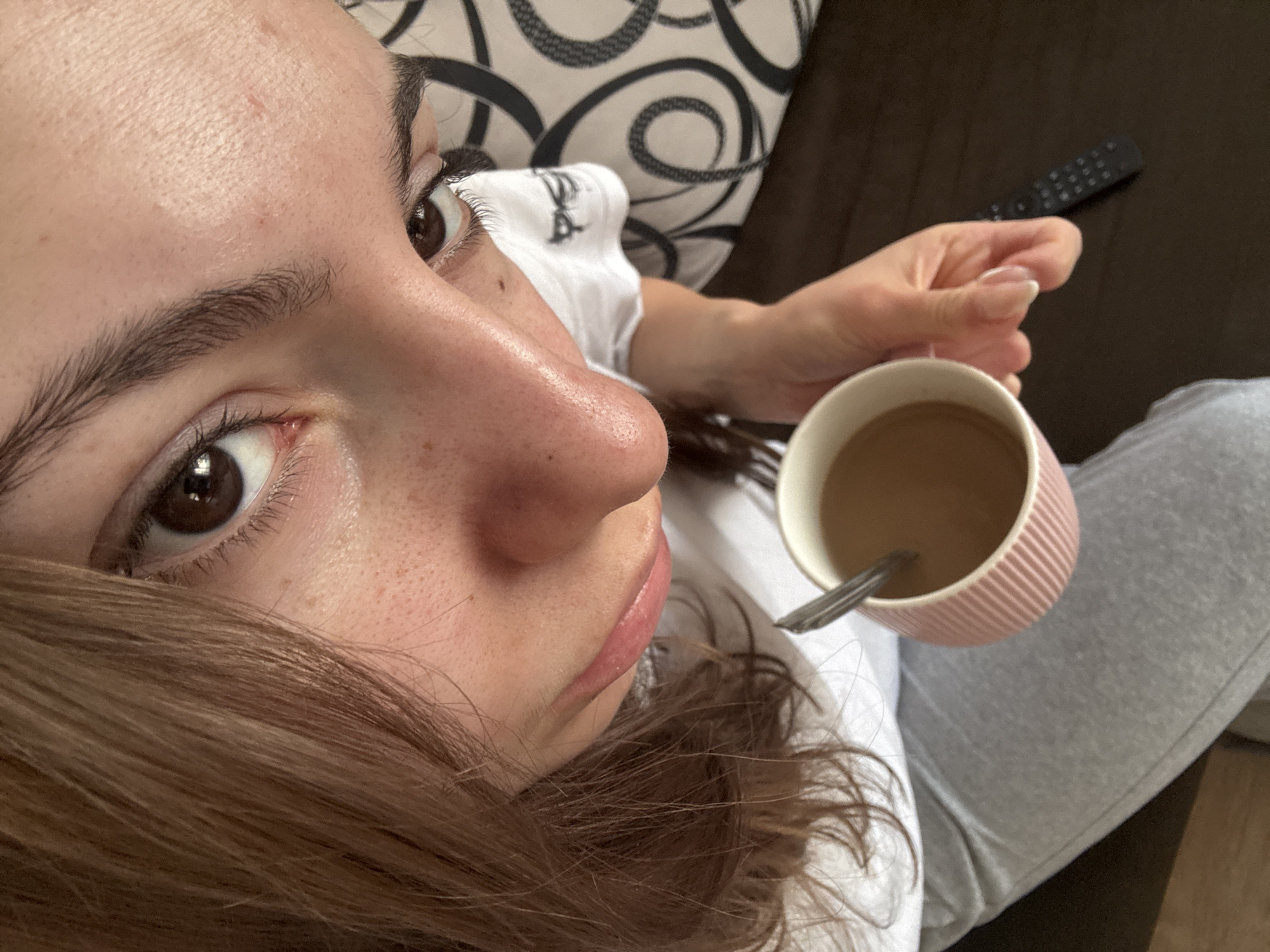 A close-up, high-angle photograph depicts a person holding a pink mug filled with a beverage. A silver spoon is visible inside the mug. The person's face is prominently featured, including their eye, eyebrow, nose, and lips. Freckles are visible on their face. The person's hair is light brown. The person is wearing a white t-shirt and gray sweatpants. A remote control is in the background, as is a cushion with a black and white pattern. The location appears to be indoors, possibly on a couch. The city and specific location are not discernible.