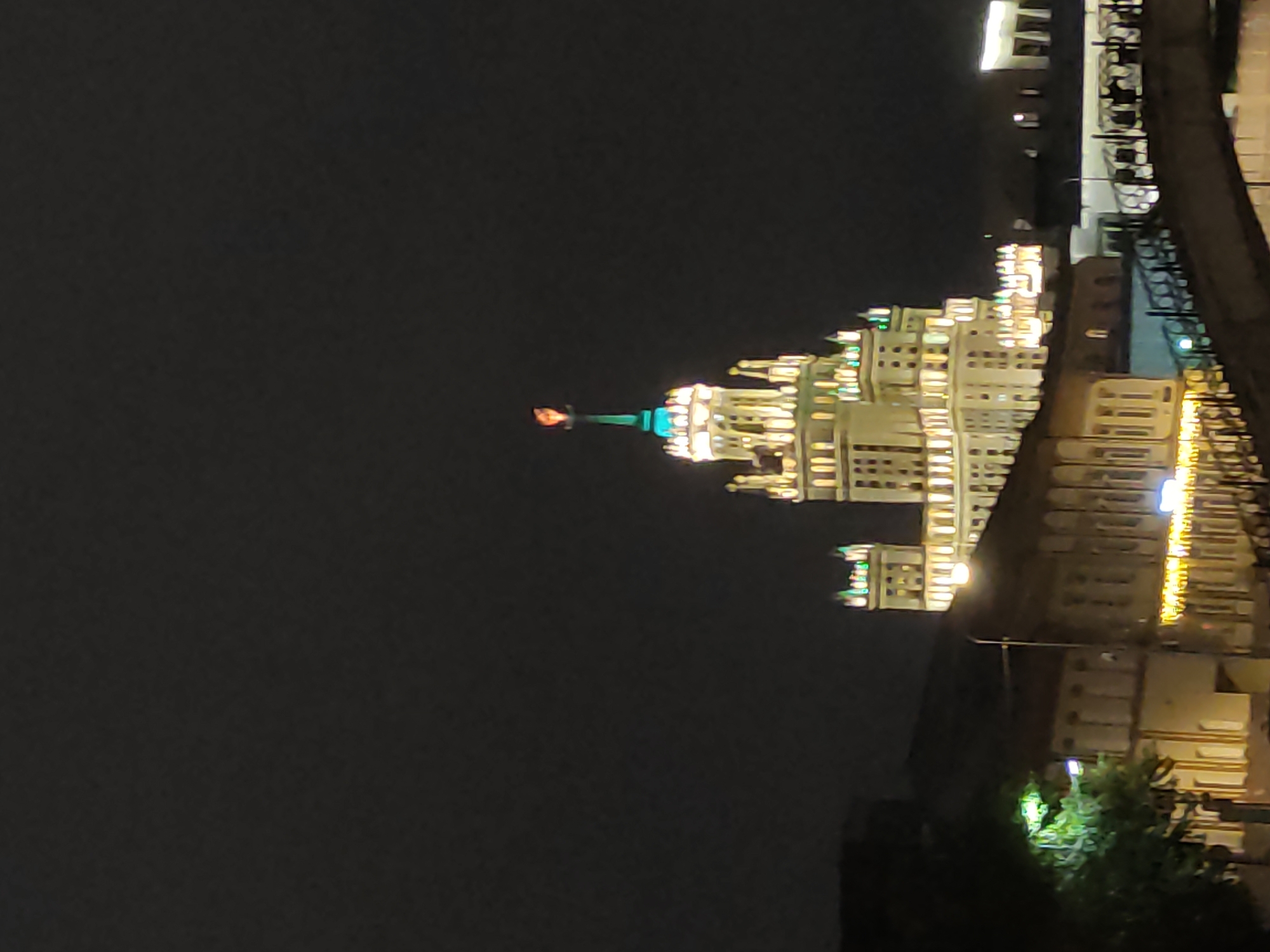 At night, an architectural structure is brightly lit. It is identifiable as one of the Seven Sisters skyscrapers in Moscow, Russia, built in the Stalinist style. At the very top of the building is an orange beacon with a spire. Additional building structures are visible, including a bridge-like structure along the upper-right. The background is dark with minimal detail. A small patch of greenery is visible in the lower right.