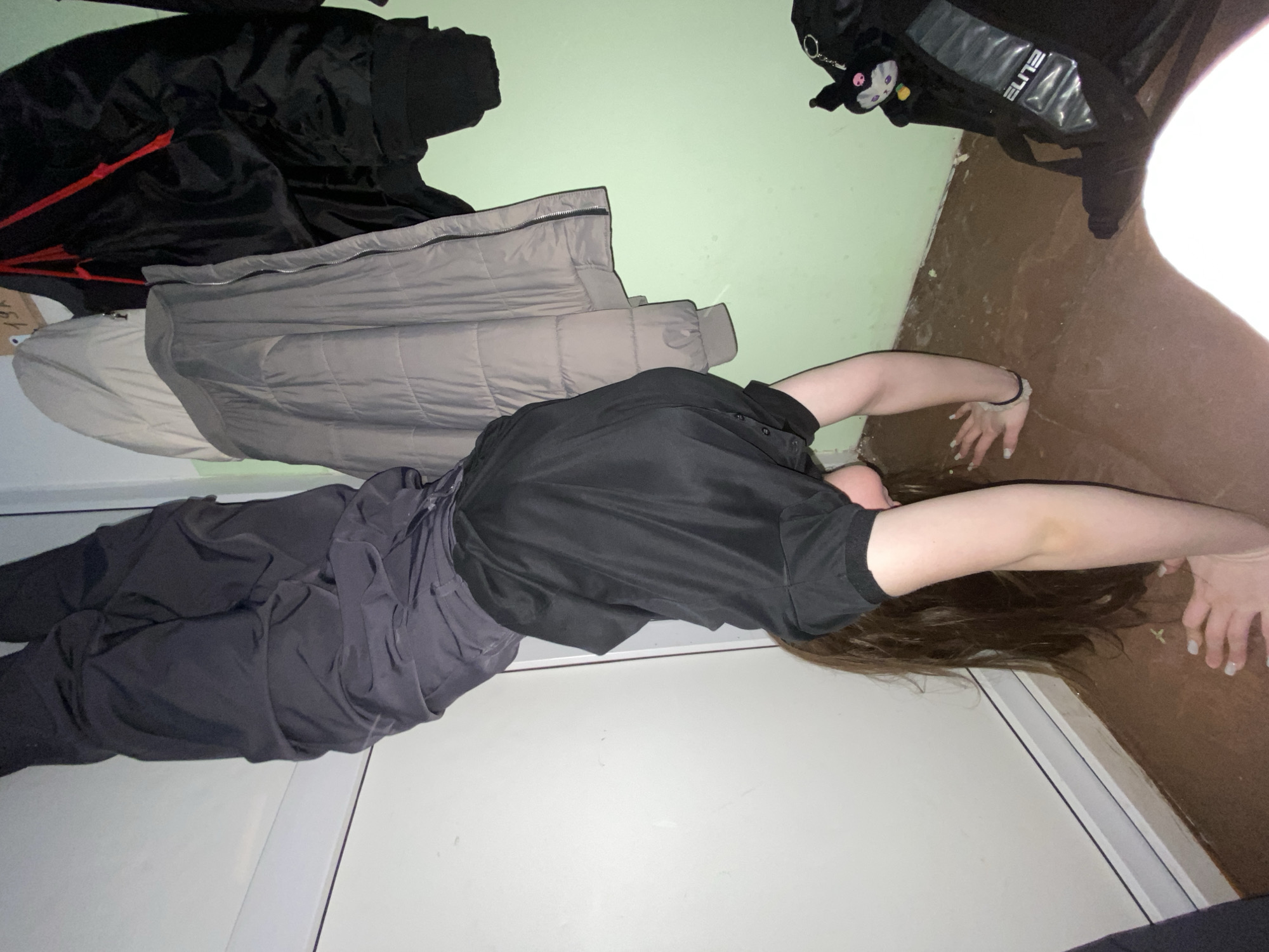 A person is lying on their back on a white surface, possibly a floor or a bed, with their arms stretched above their head. The person has long brown hair. They are wearing dark clothing. A black jacket, a gray jacket, and a black backpack are nearby against a light green wall. The floor is brown where the person's hands rest. The scene is inside a room. There is no definitive information to establish the location as Chaikovsky, Russia.