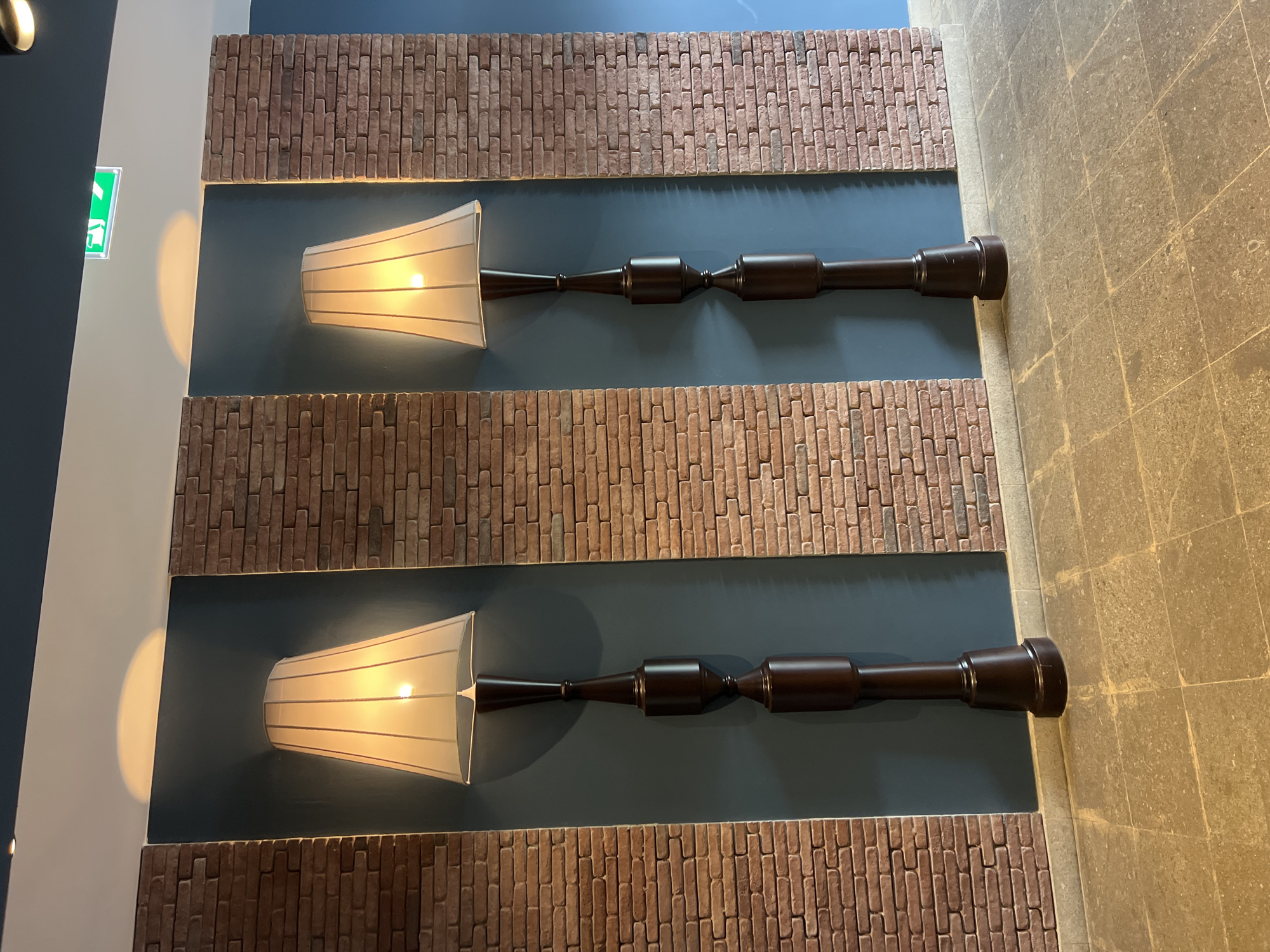 The image depicts an interior hallway with two tall, dark brown floor lamps with white, conical lampshades. The lamps are positioned in recessed alcoves with dark blue walls, flanked by sections of exposed brickwork. The floor is tiled with large, square, light-colored stones. Above the alcoves, the ceiling is dark blue, with a white section where a green, illuminated exit sign is visible in the upper right corner. The overall lighting is warm, emanating from the lamps. The scene appears to be in a public or commercial space, possibly a hotel or office building. The location is identified as Tbilisi, Georgia.
