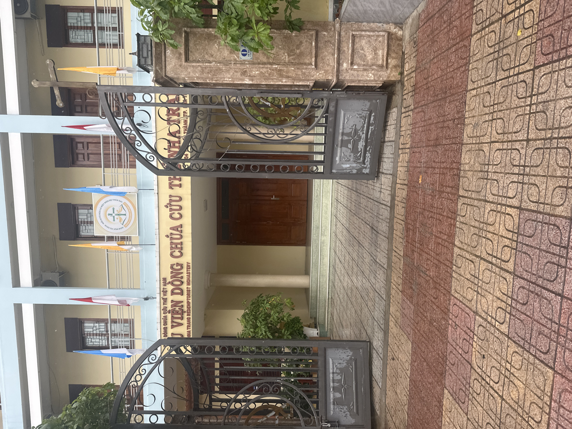 The image shows the open gates of the Nha Trang Redemptorist Monastery, located in Nha Trang, Vietnam. The monastery's façade, featuring a yellow wall with brown lettering in Vietnamese, is visible behind the gates. There are three stories, and some windows are visible in the background. A cross and various flags are visible on the left side of the building. The gates are made of dark metal with decorative circles and vertical bars. The ground is tiled with a pattern of interlocking shapes in reddish-brown and beige. The photo is taken at eye level.