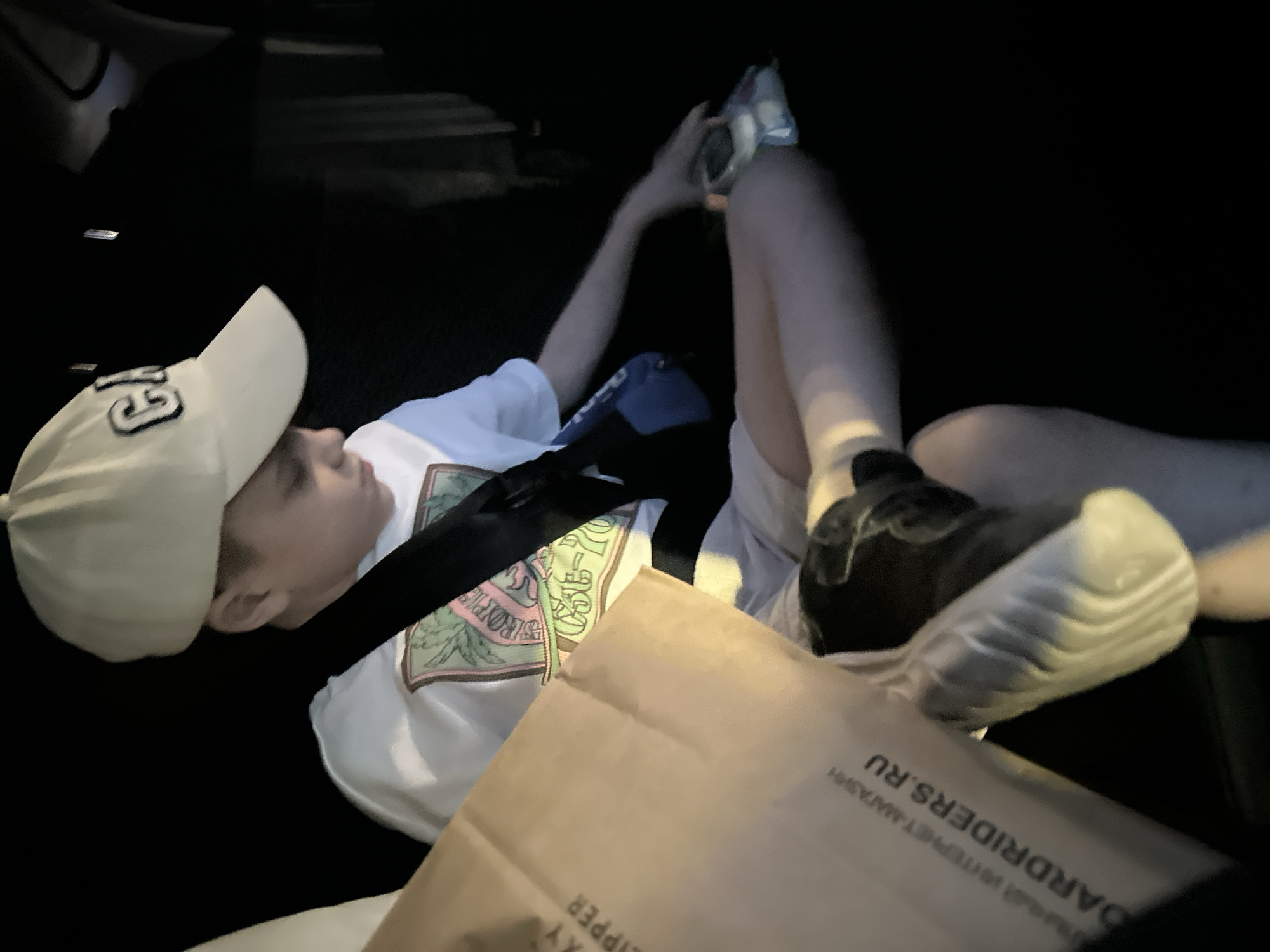 A child wearing a white t-shirt, a white baseball cap with the letter "G" on it, and black shoes, is reclined in a vehicle, secured with a seatbelt. He is holding a plastic package in his left hand. A brown paper bag with the words "OARDERIDERS.RU" partially visible on it is positioned near his feet. The image was taken in Perm, Russia, as the text on the bag indicates a Russian website. The setting appears to be inside a vehicle, possibly at night or in a dimly lit area.