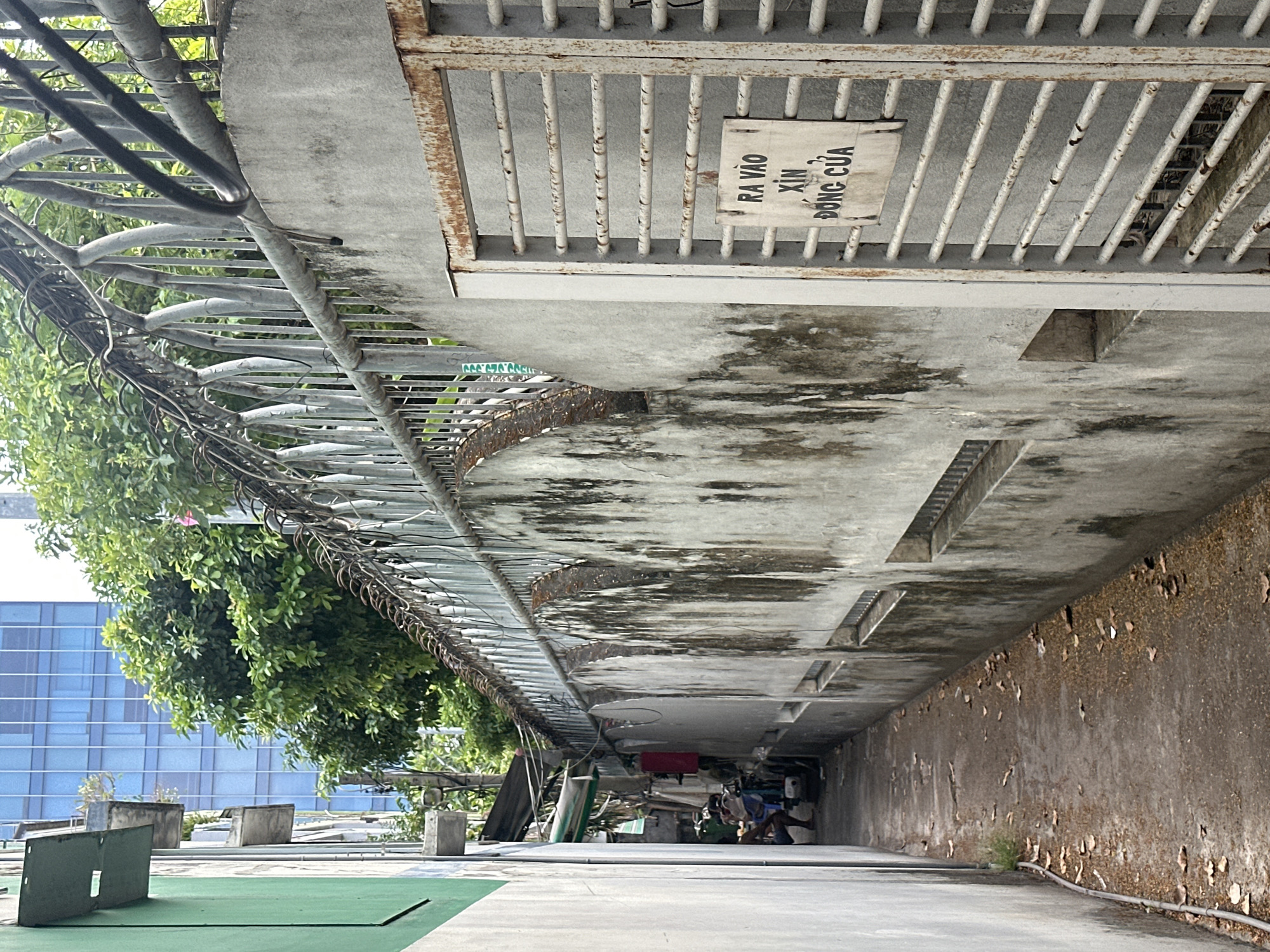 The image depicts a narrow, outdoor alleyway in Da Nang, Vietnam. On the right side of the alley, a weathered, rusty metal gate is partially open, revealing glimpses of what appears to be a fenced-off area behind it. A sign with Vietnamese text, "RA VÀO XIN ĐÓNG CỬA," which translates to "Please close the door when entering and exiting," is affixed to the gate. The wall adjacent to the gate is constructed of concrete and shows significant signs of age and staining, with dark vertical streaks. A corrugated metal fence with barbed wire runs along the top of this wall, with numerous black electrical wires tangled and draped across it.
The alley floor is paved and covered with a scattering of dry leaves and debris. On the left side of the alley, a plain, light-colored wall rises, partially obscuring the view of a green-painted structure. Further down the alley, a person is partially visible, seated near a parked scooter. Trees with lush green foliage are seen growing over the wall in the background, contrasting with the urban decay. In the far distance, a modern blue glass skyscraper is visible, suggesting the alley is located in a built-up urban area of Da Nang.