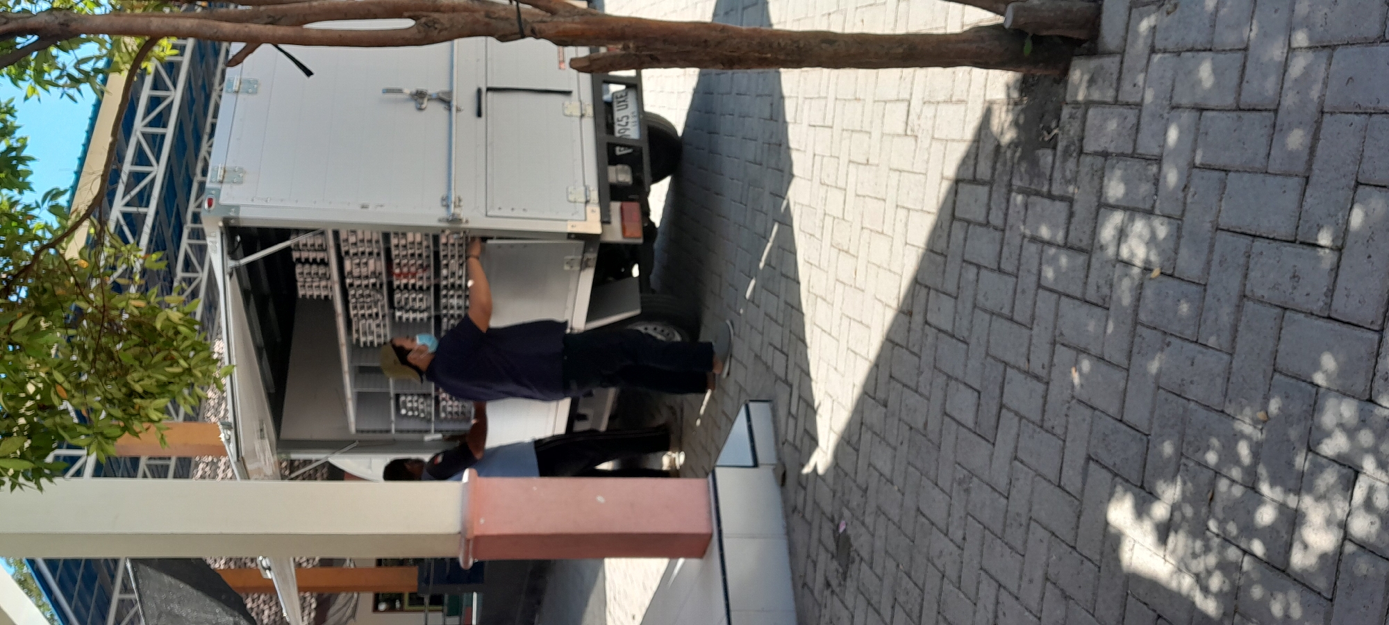 In Lemahbang Dewo, Indonesia, two men are observed near the rear of a white truck with its cargo doors open. The man in the foreground, wearing a dark blue shirt, dark pants, a light-colored cap, and a light blue face mask, appears to be interacting with the truck's interior. He is facing the truck, with his hands extended towards it, suggesting he is either loading or unloading items. The second man, standing to the left and slightly behind the first, is wearing a dark blue shirt and dark pants, and his hands are also near the truck. The truck is filled with stacked trays, possibly containing food items or other goods. The environment includes a paved walkway with interlocking grey bricks, and a portion of a white column with a pink accent is visible on the left. A tree trunk is present on the right side of the image. The background shows a blue-tiled roof structure supported by a white metal frame. The scene is illuminated by sunlight, casting shadows on the ground. A license plate on the truck reads "F 945 UXE".