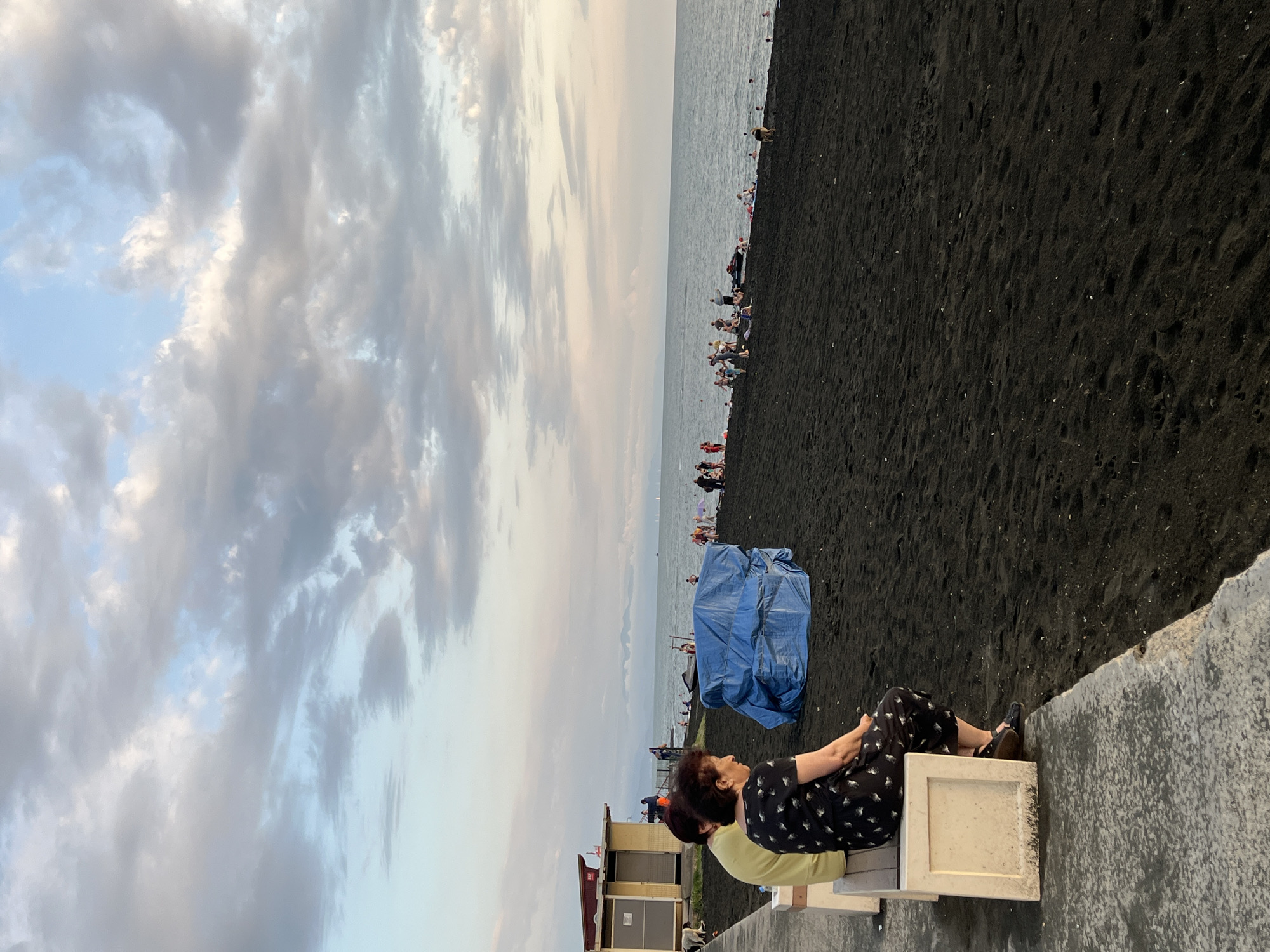 The image depicts a beach scene with a dark sand shore. The sky is filled with dramatic, cloudy formations, suggesting it may be late afternoon or evening. The sea is visible in the background, with several people scattered along the beach and in the water, implying a leisurely day at the seaside.
In the foreground, two individuals are seated on a bench facing away from the camera, looking out towards the water. The person on the right is wearing a black dress with a white pattern and black shoes. The person next to them is wearing a yellow shirt. Further down the beach, a large blue tarp covers an object or structure, possibly for shade or storage.
The overall atmosphere seems relaxed and peaceful, with people enjoying the beach. The dark sand is a distinctive feature of the location. There is no visible text in the image. The scene appears to be set in Ureki, Georgia, a location known for its magnetic black sand beaches.