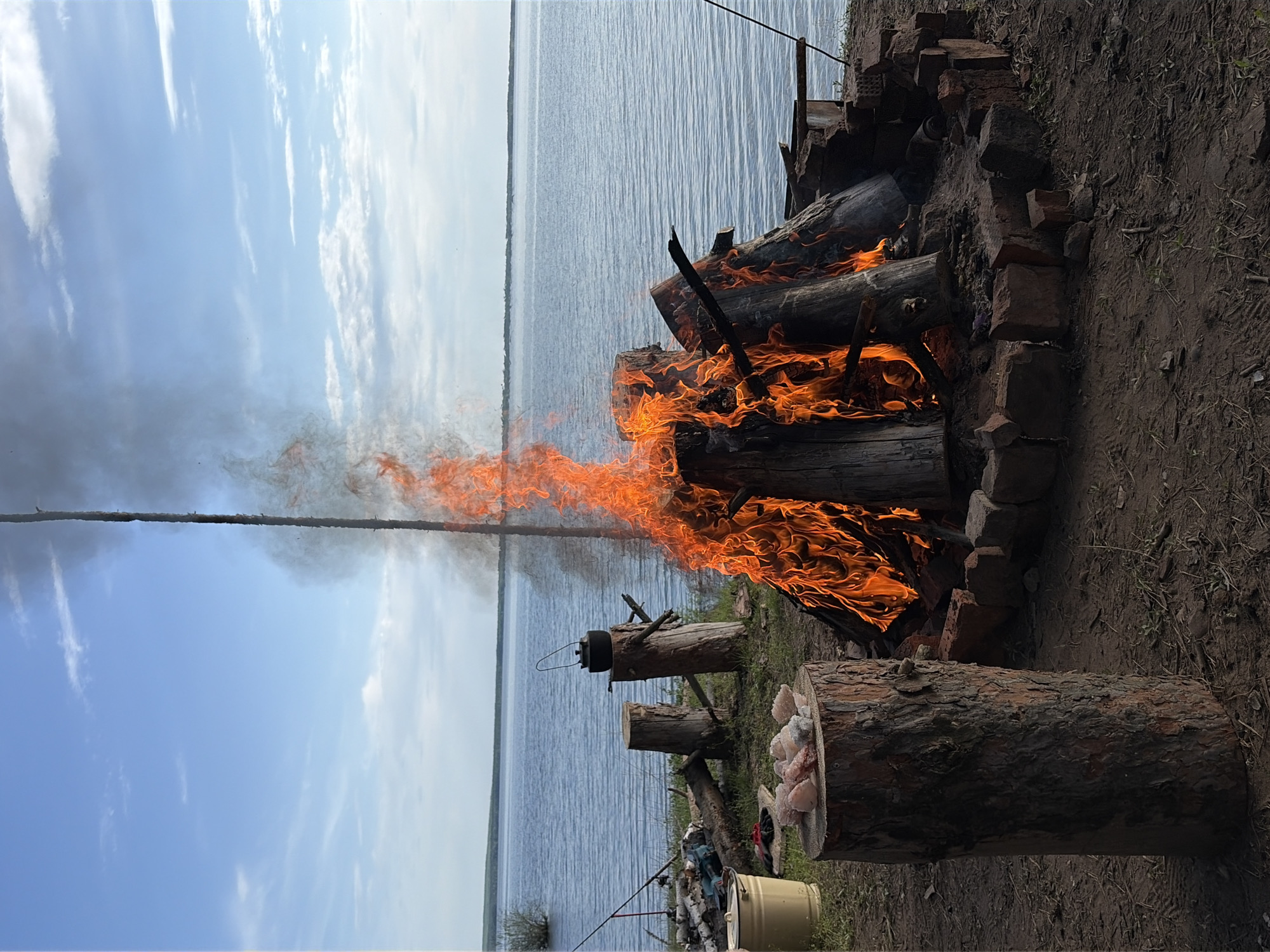 The image shows a campfire burning beside a body of water, likely a lake or river. The campfire is contained within a circle of bricks. Logs are stacked within the fire, and flames are visible. A metal pot is hanging near the fire. A log, likely for sitting on, sits next to the fire. There is a bucket on the ground. Fishing rods are present. The sky is partially cloudy. The location is suggested to be near Zabegaevo, Russia, based on information given. The image is rotated, likely taken on a phone. The picture is of a campfire cooking setup next to water.