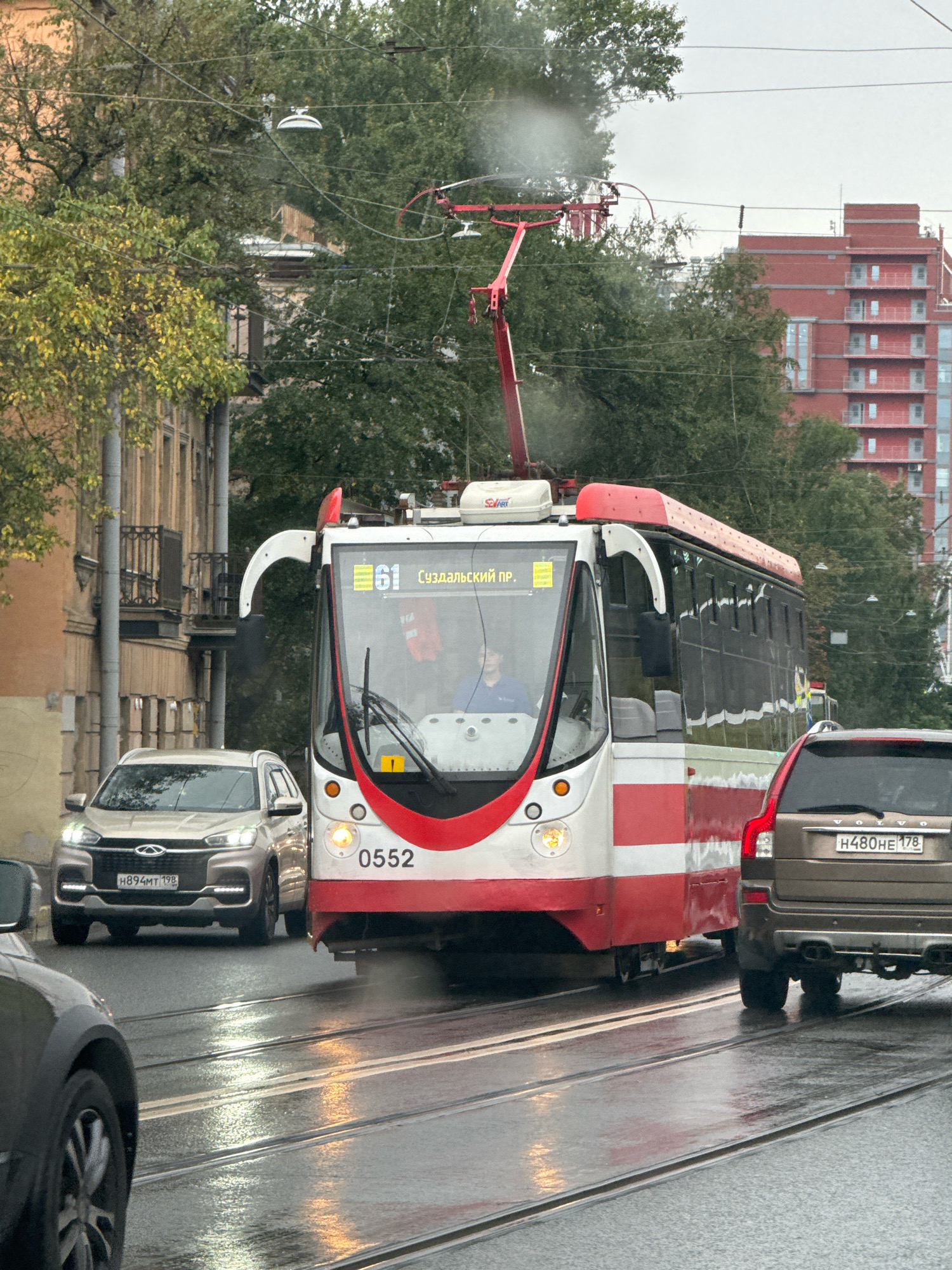 The image depicts a street scene in Saint Petersburg, Russia, on a wet, overcast day. The focal point is a red and white tram, numbered 0552, with the route number 61 and destination "Суздальский пр." displayed. The tram is on a set of tram tracks running down the middle of the street. Several cars are visible: a silver Chery SUV with license plate H894MT 198 in front of the tram, and a brown Volvo SUV with license plate H480HE 178 behind the tram. The road surface is wet, reflecting the headlights of the vehicles and the streetlights. In the background, there are trees with autumn foliage and a modern red apartment building. A person, possibly the tram driver, is visible through the tram's windshield. The scene appears to be a typical urban street with public transportation and vehicular traffic.