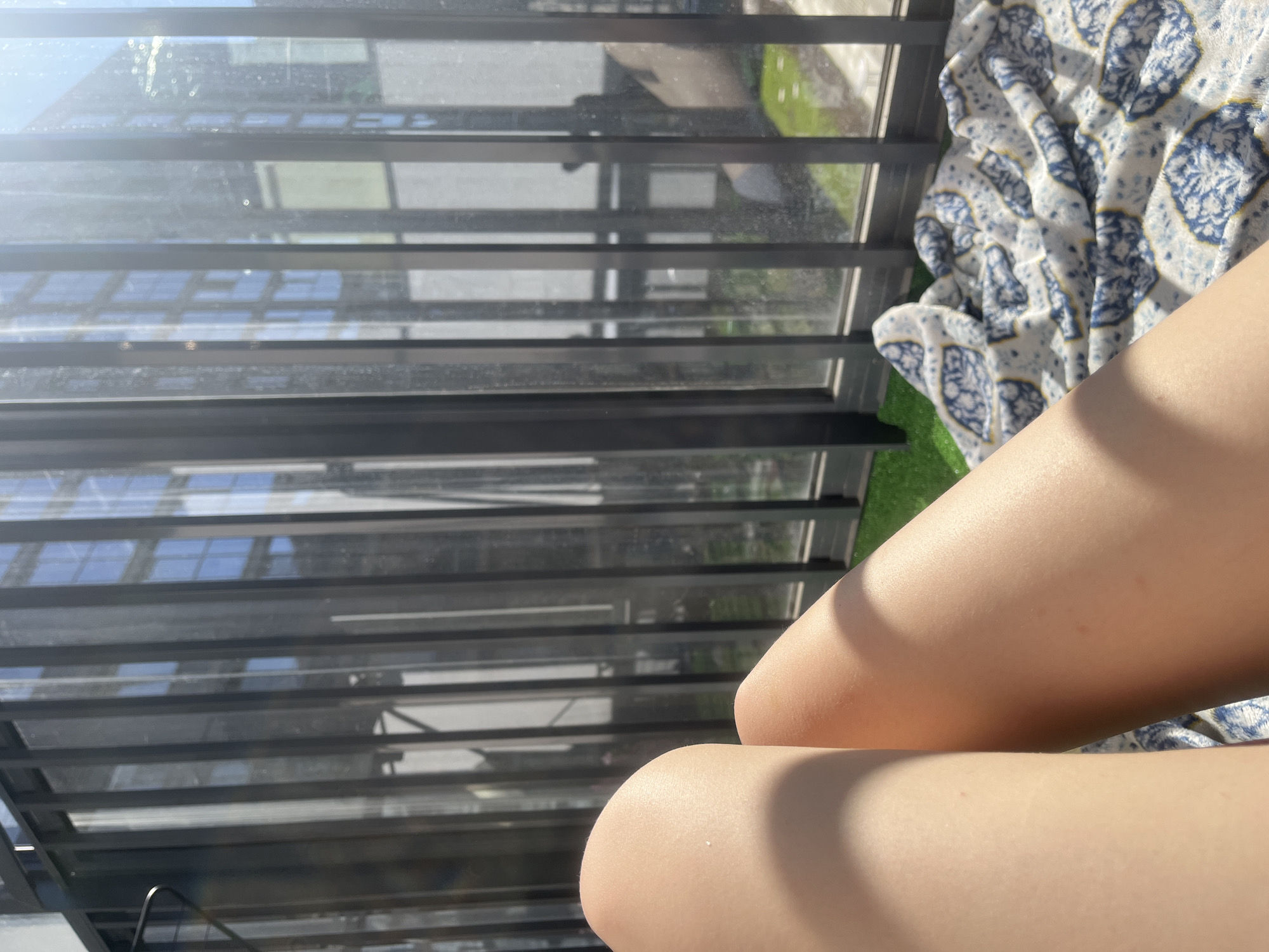 A close-up, high-angle shot shows a person's legs in the foreground, with a balcony and a view of buildings in the background. The legs are fair-skinned, and appear to be resting on a patterned blue and white blanket on what looks like artificial grass. The balcony has a metal railing with horizontal bars. Outside the balcony, there is a cityscape, possibly of Saint Petersburg, Russia. Buildings with multiple windows are visible, reflecting the sky. The light is bright and sunny.