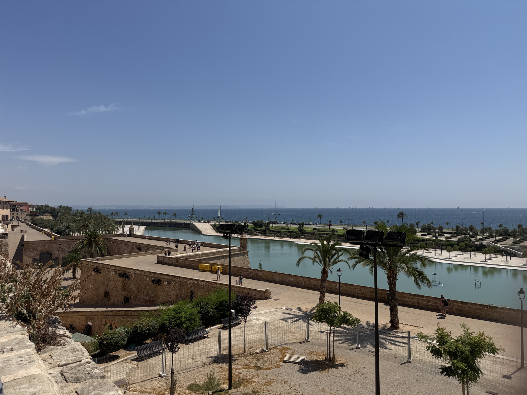 This is a wide, horizontal, eye-level shot of a waterfront area in Palma de Mallorca, Spain. The foreground is dominated by a section of a stone wall and a pathway. Beyond this, a large body of calm, turquoise water, possibly a man-made canal or harbor, stretches across the middle ground. This water feature is bordered by stone walls and lined with palm trees and manicured landscaping. Several people are walking along the pathways adjacent to the water. Further in the background, the sea is visible, dotted with numerous sailboats and other vessels. The sky is clear and blue, with a few faint clouds. Buildings, characteristic of Mediterranean architecture, are visible on the left side of the frame. The overall impression is one of a sunny, bustling urban waterfront.