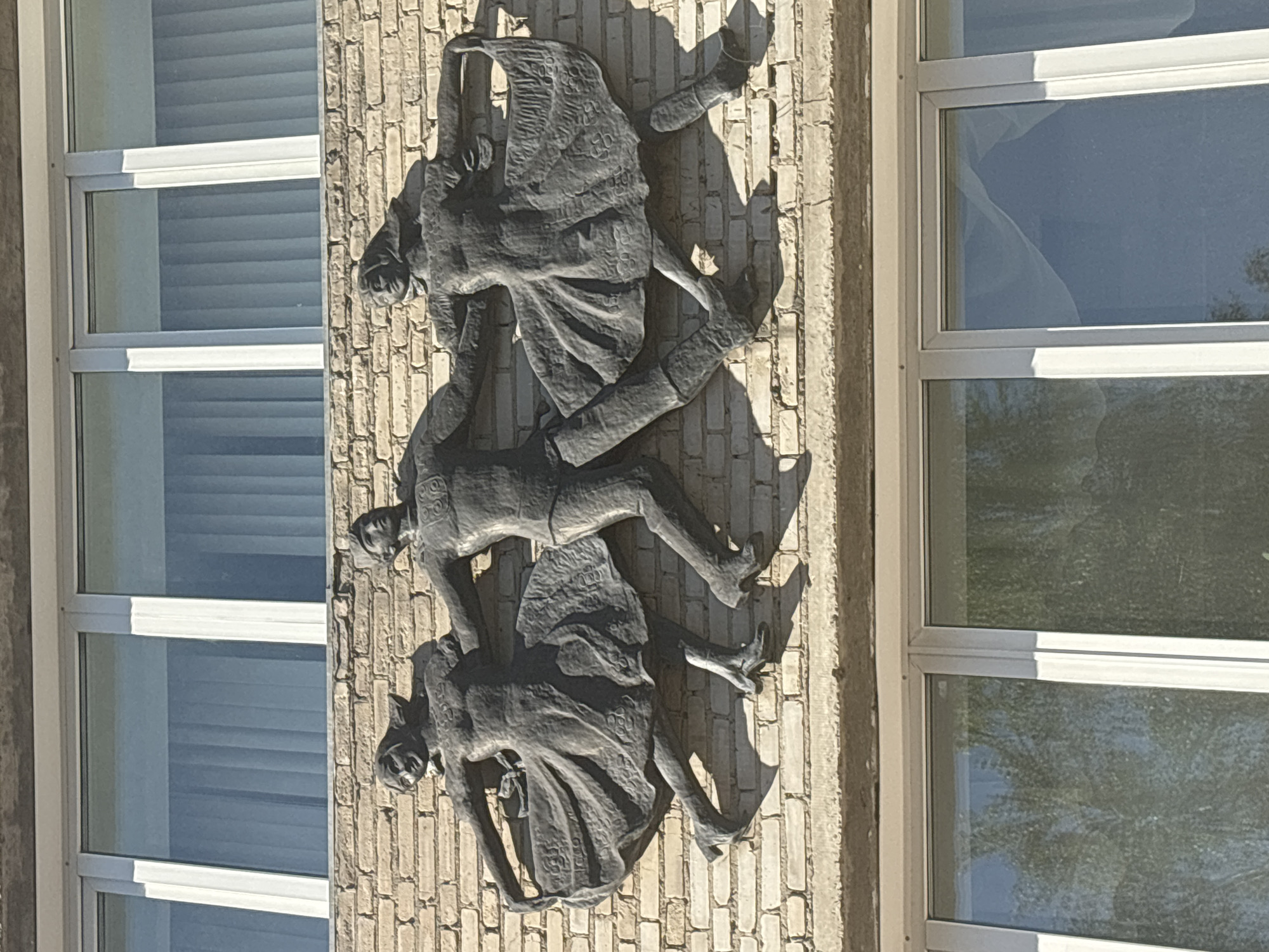 The image shows a bas-relief sculpture on the facade of a building in the city of Ob, Russia. The sculpture depicts three figures: a man in the center and two women on either side. They are shown in dynamic poses, appearing to be dancing or celebrating, with their arms linked. The figures are clad in simple, flowing garments. The man is wearing a suit, and the women are in dresses. The bas-relief is made of a dark material and is mounted on a light-colored brick wall. Above the sculpture are two sets of windows with white frames, and below it are more windows reflecting trees. The overall impression is one of movement and festivity.