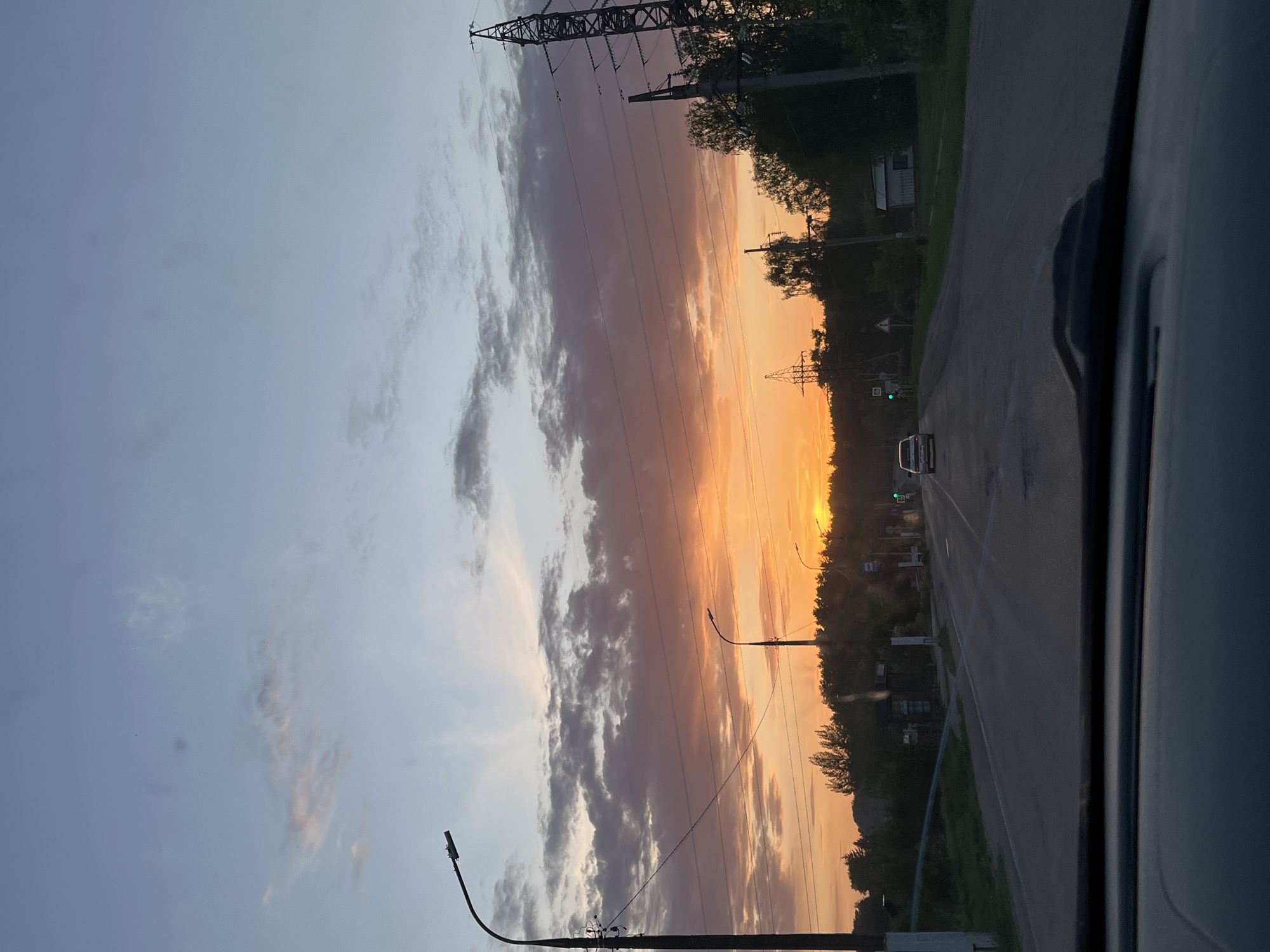Captured from inside a vehicle moving along a road in what appears to be the city of Chaikovsky, Russia, the image features a striking sunset. To the left, the sky is a mix of light blue and grey, with wisps of clouds scattered throughout. The central part of the image is dominated by a vivid orange and yellow sunset, with darker, purple-grey clouds bordering the colorful sky. Power lines and support towers are visible, intersecting the sky and adding to the industrial-urban aesthetic. The road, dark grey, leads into the distance. A light-colored car is visible on the road ahead. Trees and vegetation border the road on the right side. Street lights are also present along the roadside. The photograph appears to be taken from the passenger side of the vehicle.