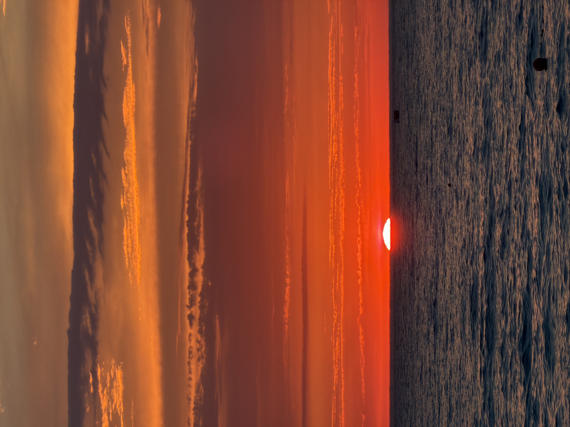 This is a vertical, eye-level, full shot of a sunset over the Black Sea in Sochi, Russia. The sun, a bright orange semi-circle, is low on the horizon, casting a warm glow across the sky. The sky is filled with streaks of orange and pink clouds, with darker clouds in the upper left quadrant. The sea is dark and choppy, with small whitecaps visible.
In the distance, on the water, there are a few dark, indistinct objects. These could be boats or buoys. The overall mood of the image is serene and tranquil, with a sense of the vastness of nature. The time of day is clearly sunset due to the sun's position and the warm hues in the sky. The location is confirmed to be Sochi, Russia, with the Black Sea as the body of water. There is no visible text in the image.