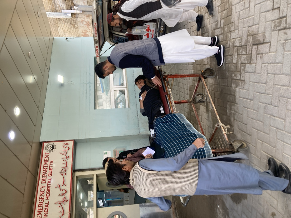 The image depicts an outdoor scene at the Emergency Department entrance of the Provincial Hospital in Quetta City Tehsil, Pakistan. A sign above the entrance clearly displays "EMERGENCY DEPARTMENT PROVINCIAL HOSPITAL QUETTA" in English and Urdu.
In the foreground, a young male patient is lying on a red-framed medical stretcher with wheels, covered from the waist down with a blue and white checkered blanket. He wears dark clothing, has a nasal cannula, and an IV line or tubing is visible near his chest. His eyes are open.
Four additional males are present. One male, wearing a dark blue shirt, gray vest, and white shalwar kameez, stands to the right of the patient, leaning over the stretcher and appearing to attend to the patient's upper body. Another male, positioned on the far right near a white ambulance with its rear door open, wears a white shalwar kameez, a dark waistcoat, and a red cap/turban. He holds an IV bag with tubing, which extends towards the patient. The ambulance bears "TOKHI FOUNDATION" and Urdu text. A third male, in the left foreground, wears a light blue shalwar kameez and a beige plaid waistcoat, facing right towards the patient. A fourth male is partially visible in the background, near the hospital entrance, holding a white paper.
The ground is paved with rectangular grey pavers. The hospital building features light blue exterior walls and glass entrance doors, with a "PHARMACY" sign visible through one door. The scene is illuminated by daylight.