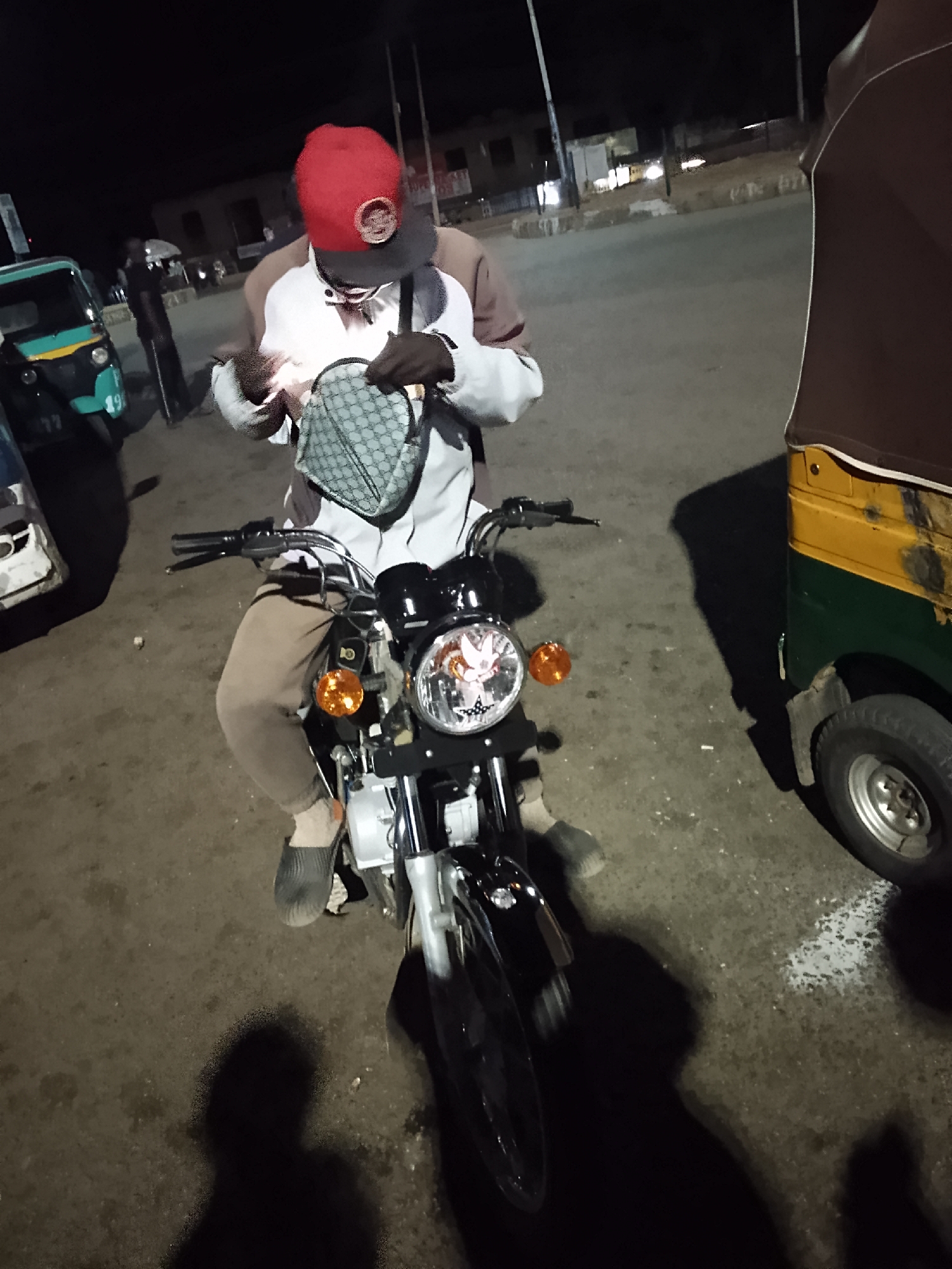 A man is sitting on a black motorcycle in what appears to be a city at night. He is wearing a red baseball cap, sunglasses, a white and brown jacket, and tan pants. He also has dark footwear that appears to be slip-on shoes. The man is holding a lighter in his left hand, while his right hand is reaching into a small, grey patterned bag that is hanging in front of him. The motorcycle has a round headlight and orange turn signals. To the left of the motorcycle, there is a light teal and white vehicle, possibly a tuk-tuk or auto rickshaw. To the right of the motorcycle, there is a yellow and green auto rickshaw. The ground is dark and uneven, suggesting a paved road or parking area. The ambient light suggests it is nighttime. There are other buildings and lights in the background, further indicating a city setting.