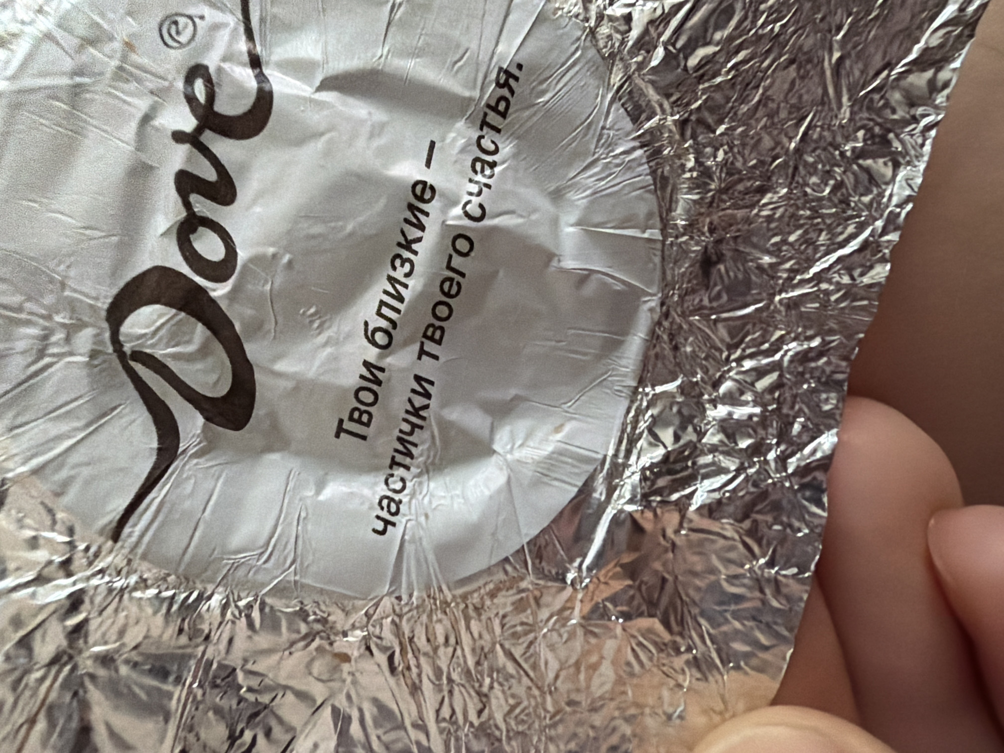 The image shows a partially unwrapped Dove chocolate candy wrapper. The wrapper is silver-colored and crumpled. The word "Dove" is printed in brown on the wrapper, along with text in Russian that translates to "Your loved ones - particles of your happiness." Part of a human hand is visible in the lower right corner of the image. The image does not provide any information to determine the location in the city of Pervomaysky, Russia.