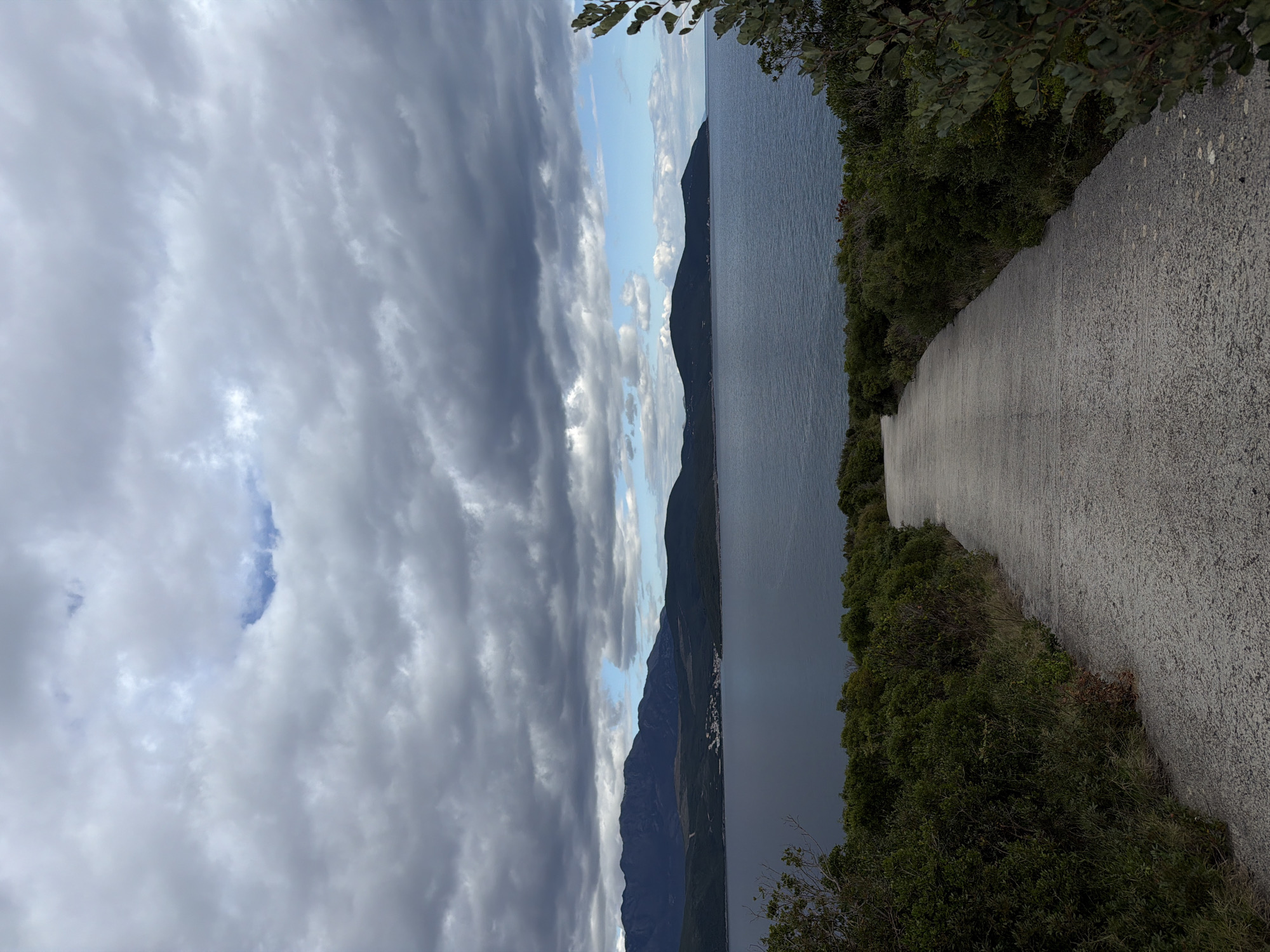 A path is adjacent to vegetation on one side, and the open sea with a mountainous coast is visible on the other. The sky is overcast with gray clouds, but there are patches of blue. The location is Radovići, Montenegro.