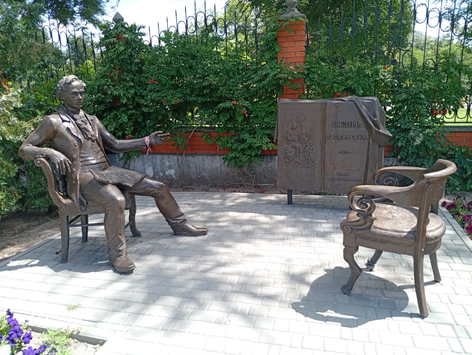 A bronze sculpture depicts a man sitting on a chair. He is wearing a suit with a jacket, vest, and shirt with a cravat. His right arm is extended outward, with his index finger pointing. The sculpture is located outdoors on a paved area with gray square tiles. Behind the sculpture is a brick wall with a decorative black metal fence on top, and lush green bushes. To the right of the main sculpture, there is another bronze sculpture of an empty chair. Next to the seated man's sculpture, there is a large bronze book sculpture. The book has a cover with an embossed image and text, including the title "КОБЗАРЬ" and the name "Т. ШЕВЧЕНКА". There is a shadow cast by the sculpture of the seated man on the tiled ground. In the foreground, to the left, there are purple flowers in a planter. The lighting suggests it is daytime, with sunlight casting shadows.