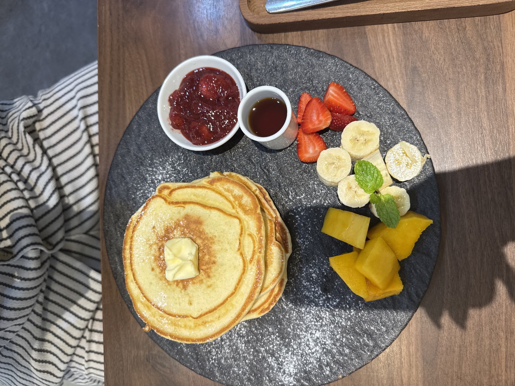 A top-down shot shows a plate of food on a wooden table, likely in Đà Nẵng, Vietnam, given the style of food. The plate, a dark, possibly slate-like surface, holds a stack of pancakes topped with a pat of butter and dusted with powdered sugar. Alongside the pancakes, there are portions of fresh fruit, including sliced strawberries, bananas, and mango cubes. Two small white bowls contain strawberry jam and a liquid, likely syrup. A sprig of mint rests among the banana slices. To the left of the plate, part of a striped fabric is visible, suggesting a tablecloth or article of clothing. The table surface is made of wood with a dark, consistent grain. The lighting suggests a bright, possibly sunny, day.