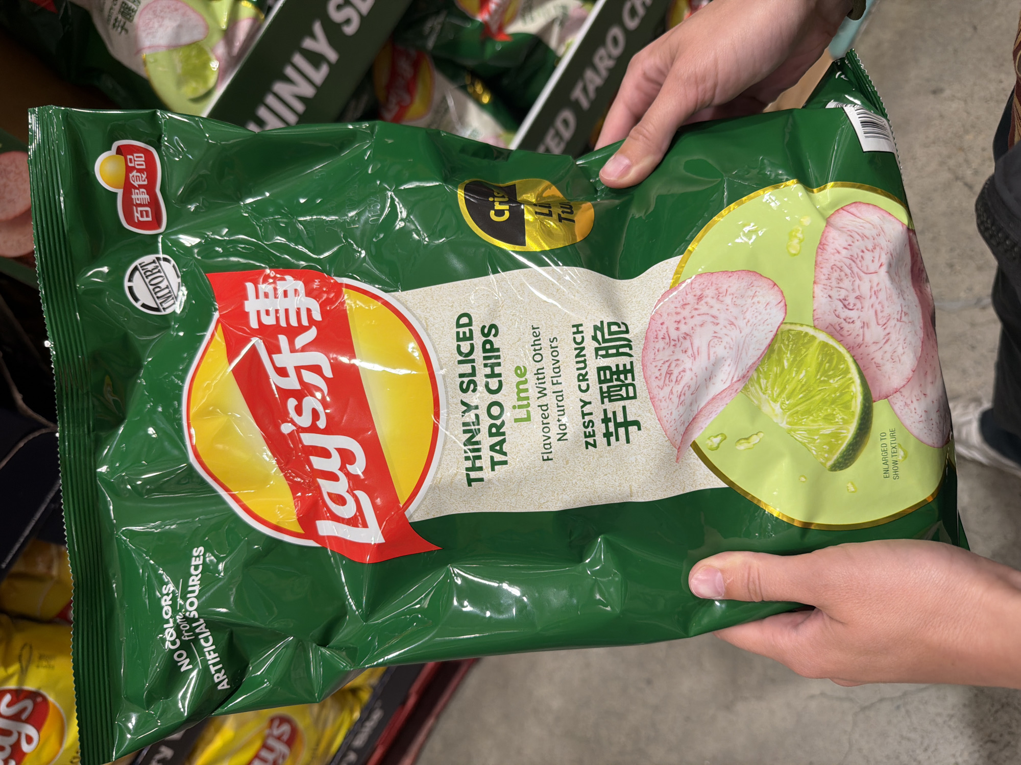 A person's hands hold a large, crinkled green bag of Lay's thinly sliced taro chips flavored with lime. The bag is the primary subject, featuring the Lay's logo in red and yellow, along with Chinese text, and an illustration of lime slices and taro root. The bag also displays text indicating "NO COLORS from ARTIFICIAL SOURCES" and "Zesty Crunch." In the background, other bags of chips are visible, some yellow and others green with "EINLY SE" visible on one. The environment appears to be a retail setting, possibly a grocery store aisle, with a concrete floor. This scene is located in Beaverton, Oregon, United States of America.