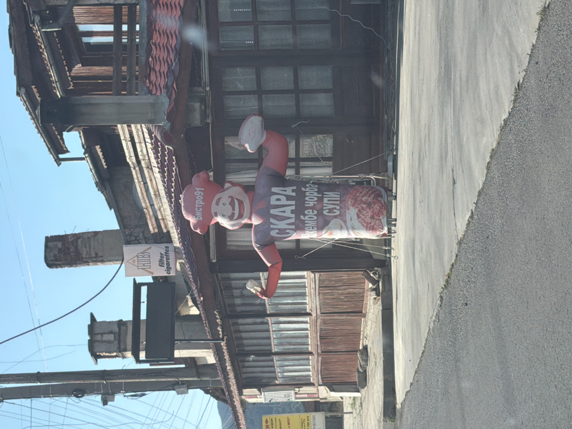 In Kamchiya, Bulgaria, a scene unfolds on a sunny day, featuring an outdoor setting in front of a building with a red-tiled roof and wooden facade. Dominating the foreground is a large, inflatable character dressed as a chef, with a red chef's hat and a smiling face. The character's torso is adorned with text in Cyrillic script, including "СКAРА" (SKARA), "джембе чорба" (Dzembe Chorba - a type of soup), and "СУПИ" (SOUP). The inflatable figure is holding a white object in its right hand. To the left of the inflatable chef, a sign with a triangular logo and the text "filter cigarettes" is visible. The building behind the character appears to be a restaurant or bistro, identified by a sign reading "Бистро91" (Bistro91) on the inflatable. The environment includes a paved area, a road, and utility poles with numerous wires. The sky is clear and blue.