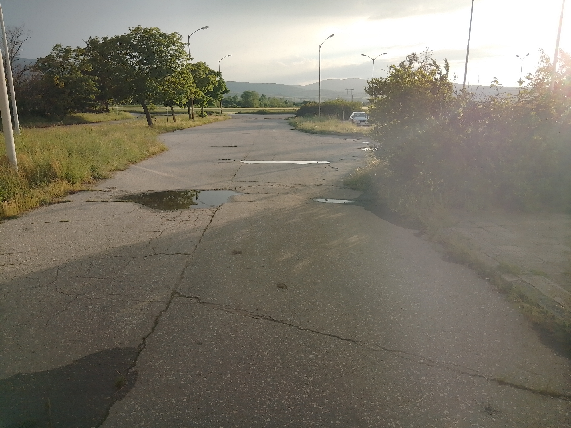 The image captures an outdoor scene featuring a deteriorating paved road or lot under a bright, possibly late afternoon or early morning sky. The asphalt surface is heavily cracked and features several large potholes, some of which contain standing water, reflecting the sky. Long shadows stretch across the ground from the left side, indicating the sun is low in the sky, either rising or setting. A strong lens flare is visible on the right side of the image, suggesting the sun's position.
Along the edges of the paved area, there is abundant, overgrown grass and wild vegetation. Several trees with lush green foliage line the left side of the road, and more scattered trees and bushes are visible further back. Multiple tall streetlights, characteristic of those found along roads or in parking areas, are placed at intervals on both sides, extending into the background.
In the mid-distance on the right, partially obscured by green foliage, a light-colored (possibly white or silver) car is parked. Beyond the immediate surroundings, a landscape of rolling hills or mountains can be seen under a partly cloudy sky, extending to the horizon. The scene appears deserted, with no people visible. The cracked pavement, potholes, and untamed vegetation suggest a place that is disused, neglected, or in a state of disrepair. No text is visible in the image.