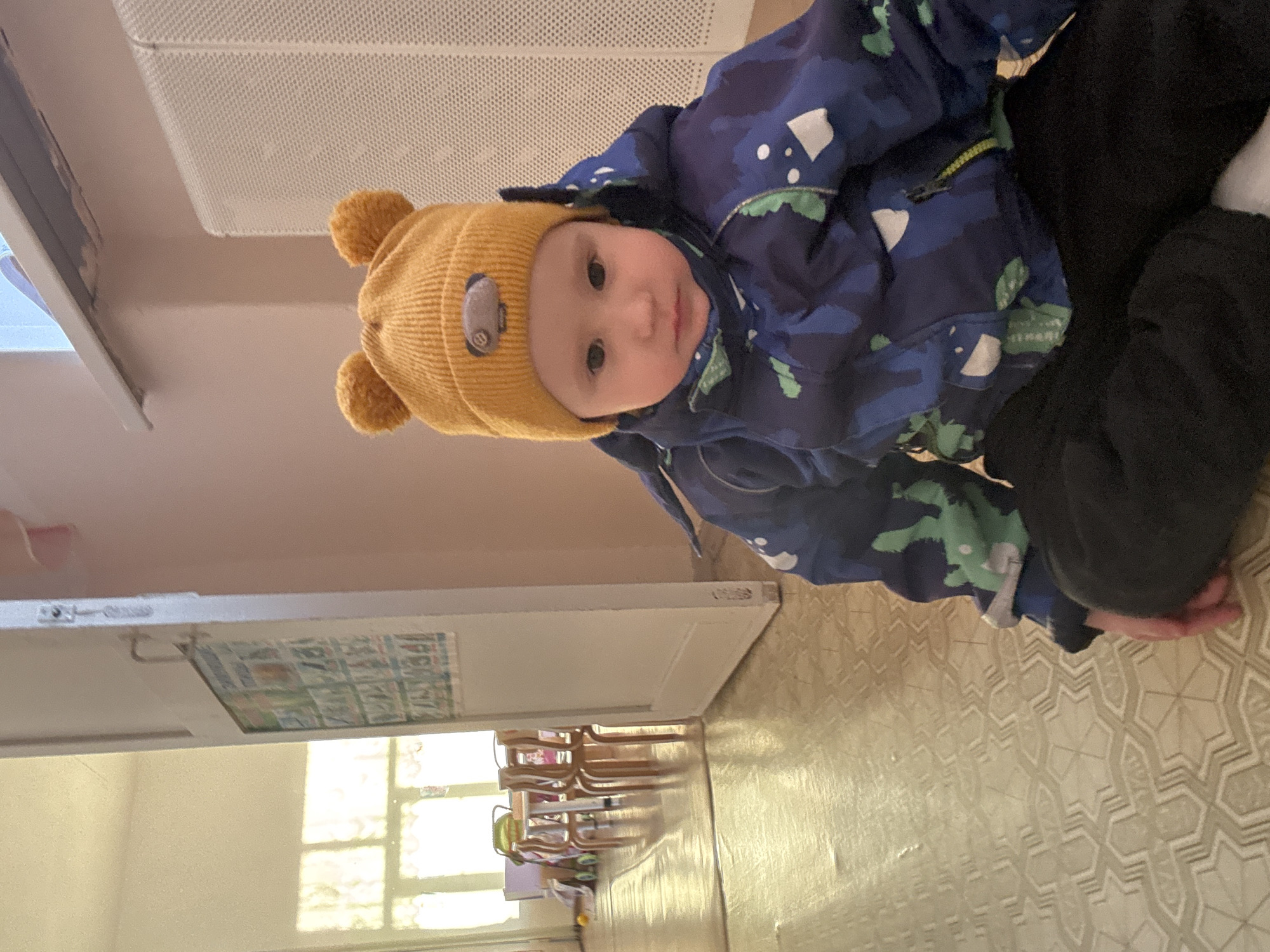 A toddler wearing a yellow hat with bear ears and a blue camouflage jacket is positioned at the top of the image. The child has fair skin and is looking at the camera. The toddler is wearing black pants. The background includes a room with beige walls, a tiled floor, and a radiator on the wall. A doorway leads to a room with children's furniture, including tables and chairs. Based on the style and contents of the room, this might be a daycare or preschool setting. There is no indication in the image of the location in Noviy, Russia.