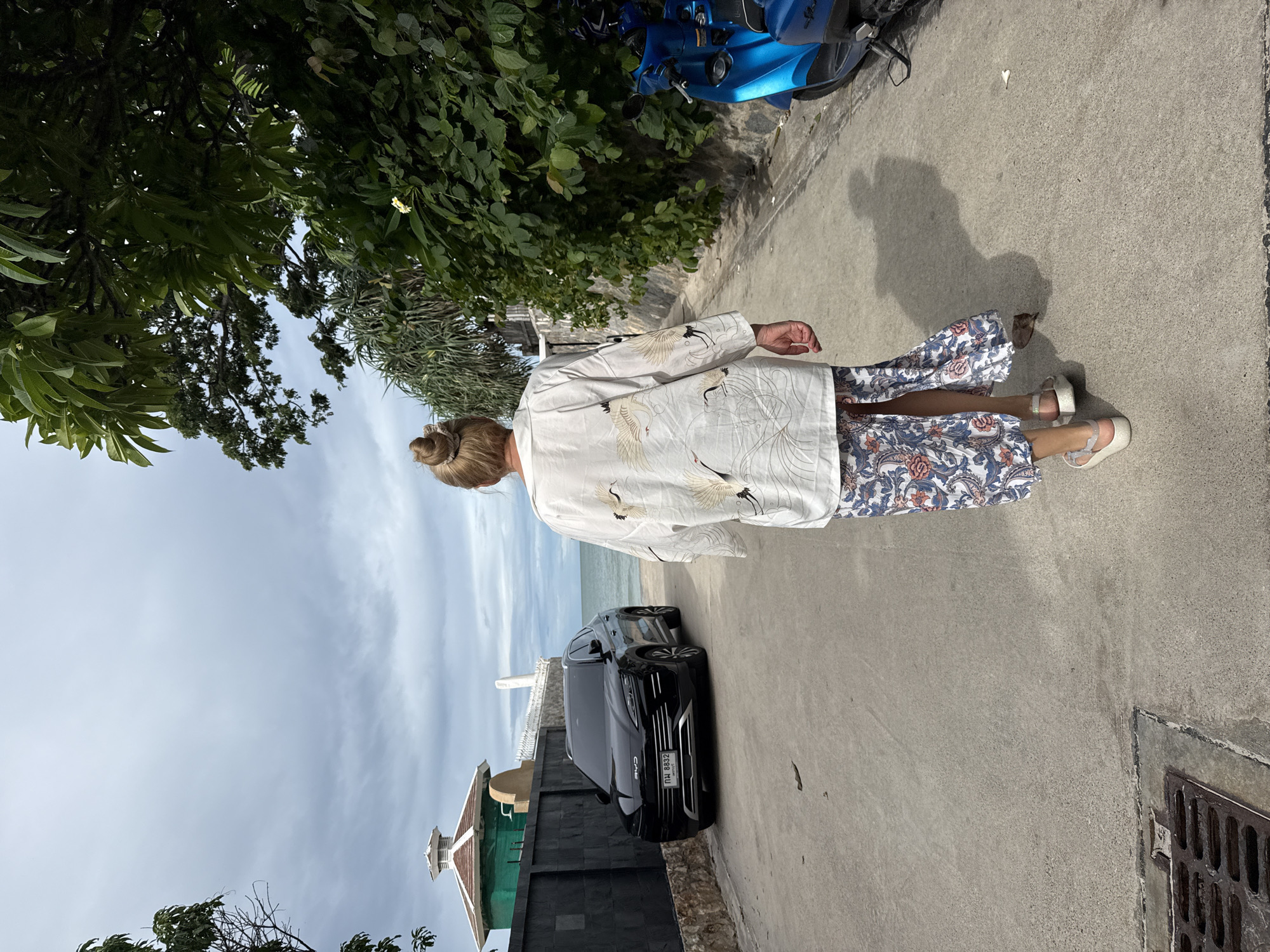 A woman with blonde hair in a bun walks away from the camera down a paved road lined with lush green foliage on the right and a dark grey wall on the left. She is wearing a white kimono-style top with a crane pattern and a long, flowing skirt with a floral pattern in blue, white, and red. She has on white sandals. In front of the wall, a black SUV with a license plate reading "กพ 8832" is parked facing the ocean. Further down the road, a bright blue scooter is parked. The scene appears to be in a coastal area, with the ocean visible in the background. The sky is overcast with gray clouds. The location is identified as a road in Hua Hin City Municipality, Thailand.