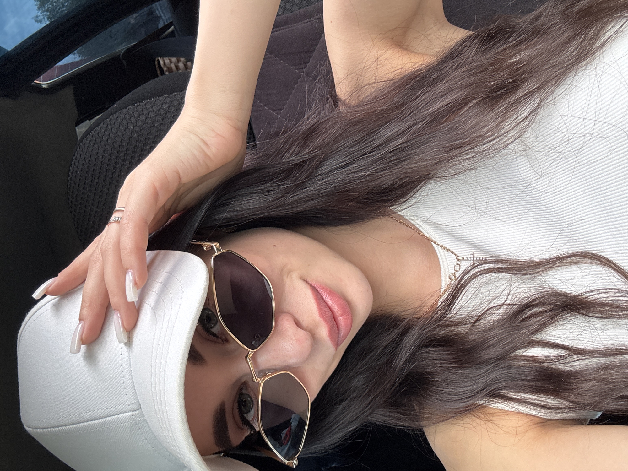 A young woman with long brown hair, wearing a white tank top and gold-framed sunglasses, poses indoors, likely in a car. She is wearing a white baseball cap, and her hand with long, white fingernails rests on the cap. A ring is visible on her finger. The car seat and the interior of the car are black. The photo does not offer definitive information about whether it was taken in the city of Chaikovsky, Russia.