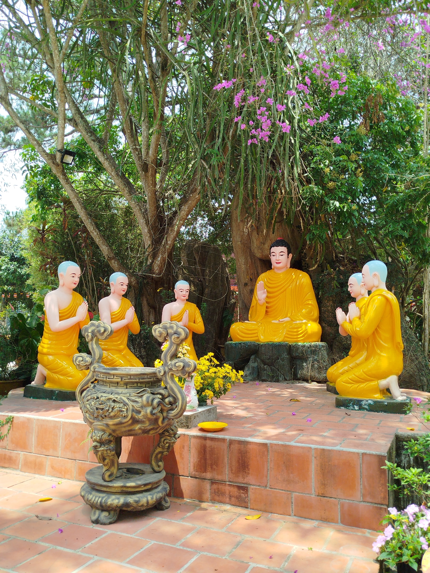 The image depicts a Buddhist shrine situated on a tiled platform outdoors. A central Buddha figure, depicted in a golden hue, sits in a meditative posture on what seems to be a rock base. Surrounding the Buddha statue are several smaller figures also painted in golden yellow, in poses of reverence. These figures appear to be monks.
A large, intricately designed, bronze incense burner sits prominently on the lower level of the platform in the foreground. A variety of lush greenery surrounds the shrine, including trees and flowering plants. The setting appears to be a garden or a park-like environment, suggested by the foliage and serene atmosphere. The presence of flowers and neatly arranged tiles suggests a well-maintained and peaceful location. The location of this shrine is in Da Lat, Vietnam.