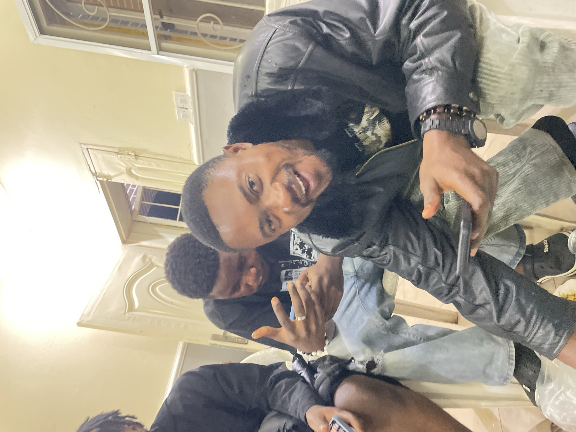 The image shows a room with three individuals. The person in the foreground is wearing a black leather jacket with fur trim and has a watch and bracelet on his left wrist. His jeans are light-colored corduroy. He is holding a phone and smiling at the camera. Another individual is partially visible behind him, wearing a black t-shirt and holding up three fingers. A third person is partially in the frame at the bottom, wearing black clothing and holding a phone. The room is painted a light color. There are windows with decorative trim visible in the background. It's impossible to verify that this picture was taken in Jos, Nigeria.