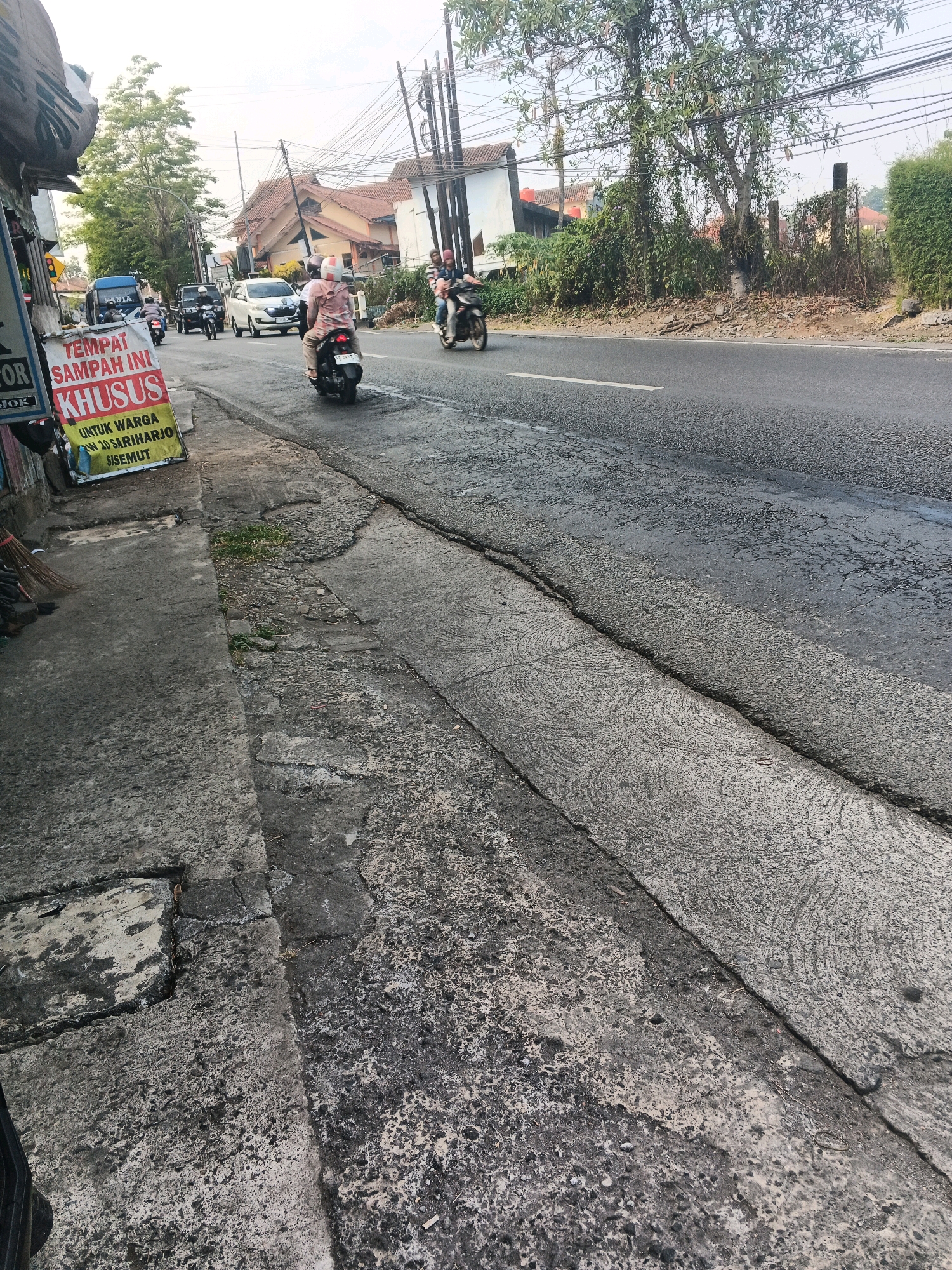 A street scene in Ungaran, Indonesia, shows a two-lane road with traffic. On the left side of the road, a sidewalk is visible, with a yellow and red sign that reads "TEMPAT SAMPAH INI KHUSUS UNTUK WARGA RW 10 SARIHARJO SISEMUT." Several motorbikes are present, with two in the foreground on the right side of the frame and one in the middle of the road. A white car is also visible in the traffic. Buildings and trees line the right side of the road, with power lines crisscrossing overhead. The road surface appears to be asphalt with some cracks and patching. The sky is overcast, suggesting a cloudy day.