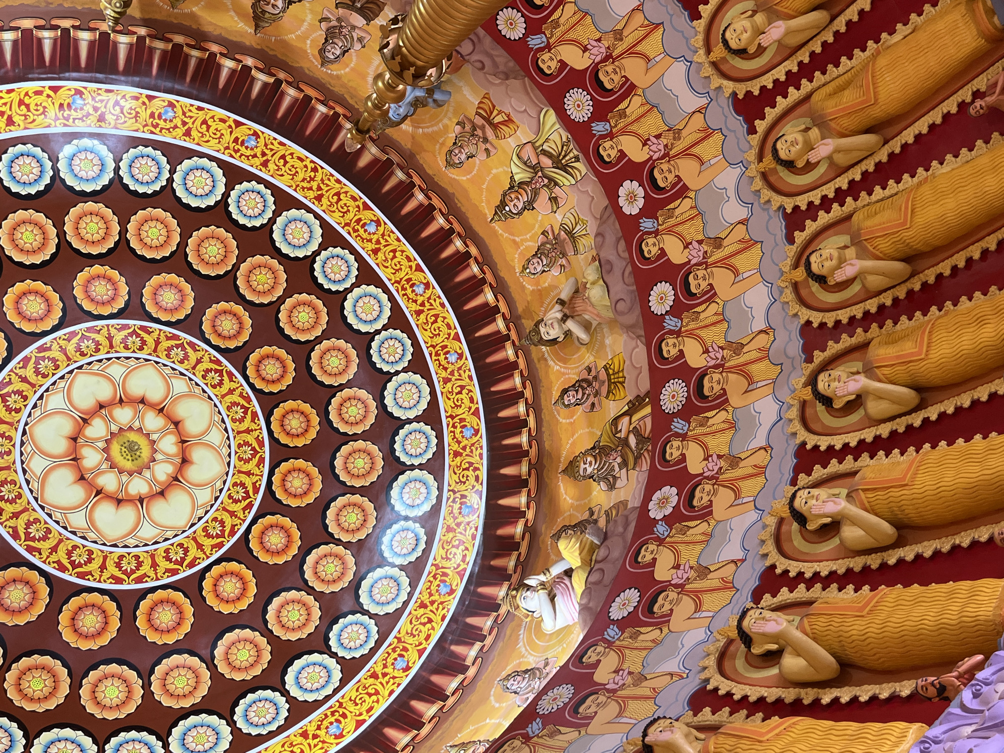 This image depicts the ornate ceiling of the Buddhist Temple in Walgama, Sri Lanka. The ceiling is elaborately decorated with a large lotus flower motif at its center, surrounded by concentric rings of smaller floral designs in shades of orange, yellow, blue, and red. The walls below are adorned with murals and statues of Buddhist figures. Rows of golden statues of individuals in monastic robes, each with a halo, are prominently displayed. Above these, a band of figures in prayer is painted against a sky-like background, interspersed with white floral designs. The entire scene is rendered in vibrant colors and intricate detail, suggesting a place of significant religious and cultural importance.