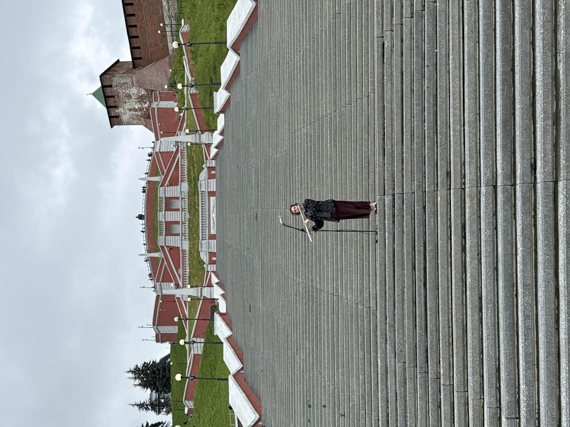 A young person stands on a wide, stone staircase, holding a flute and playing it. The person is wearing a dark jacket and maroon pants. A microphone is positioned in front of them. The staircase is very long and ascends towards a large, ornate, red and white structure with a green-topped tower visible in the background. The structure appears to be part of the Nizhny Novgorod Kremlin, a historic fortress in Nizhny Novgorod, Russia. The sky is overcast.