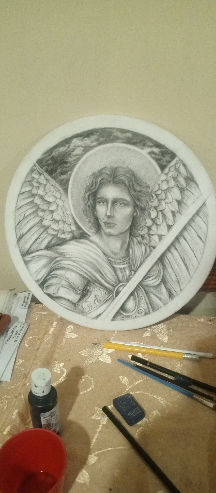 The image depicts a pencil drawing of an angel figure, possibly Archangel Michael, presented on a circular white surface. The angel has curly hair, a halo, wings, and is holding a sword. The background features cloudy, smoky shapes.
The drawing rests on a patterned gold-colored surface, likely a tablecloth or fabric. Around the drawing are art supplies, including pencils, brushes, a black ink bottle, a red cup containing liquid, and an eraser. There are also small papers with writing on them.
The location is Tinaquillo, Venezuela. The environment appears to be an indoor workspace.
