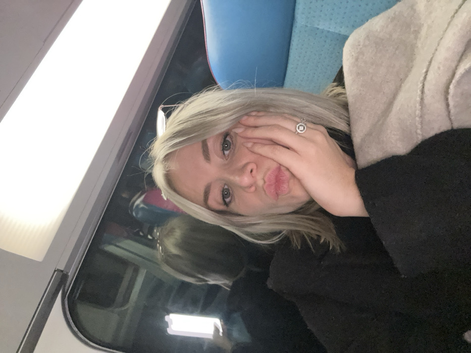 A woman with blonde hair is lying on a train seat. Her hair is blonde and slightly messy. She's wearing a black sweater and a beige jacket. She has her hand on her face, near her mouth, and is making a kissing face. She's wearing a silver ring with a large stone. The train window is behind her, with a blurry reflection of the interior. The seat appears to be blue. The overhead light is bright.