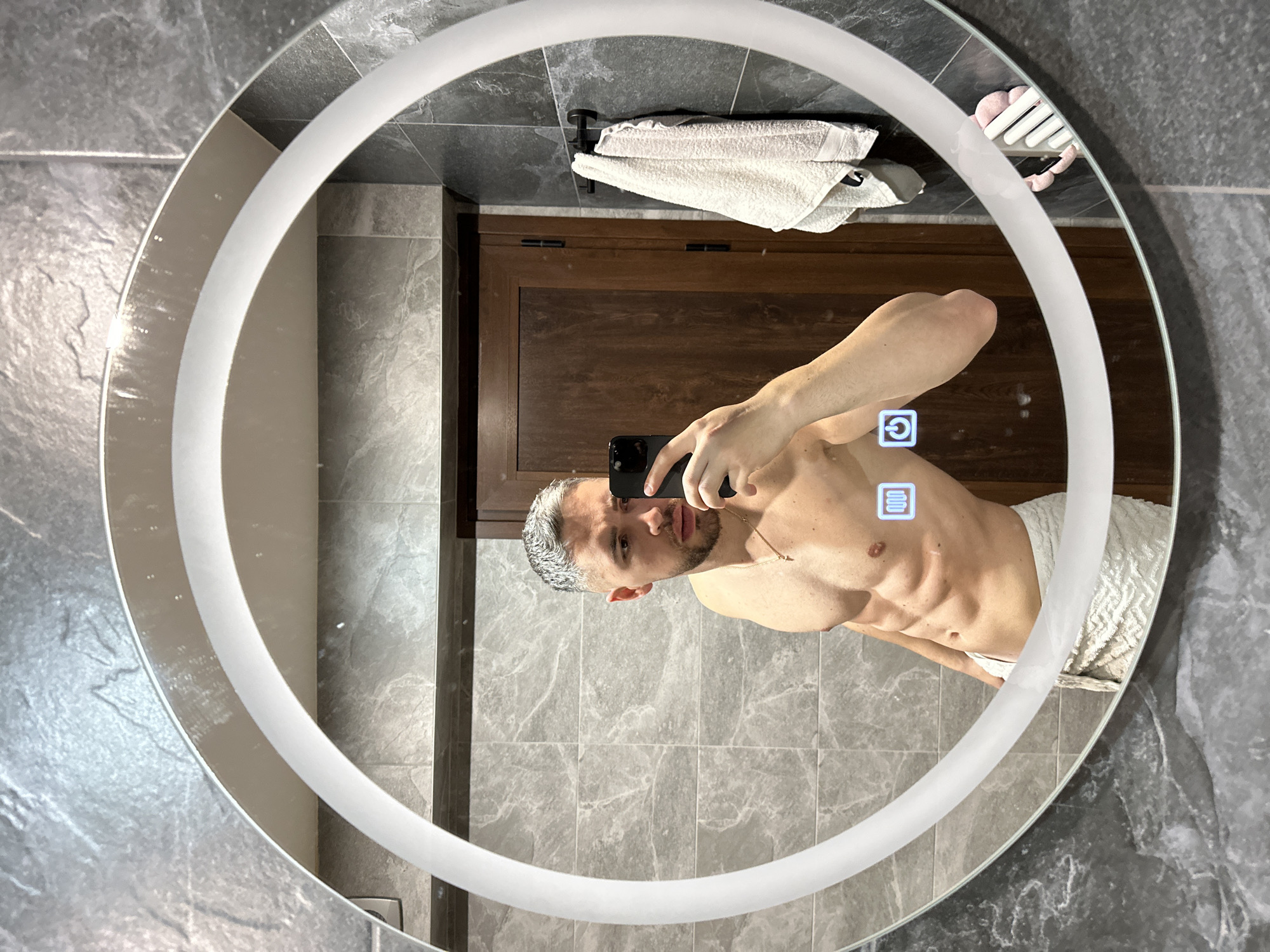 An image, rotated 90 degrees clockwise, displays a shirtless man taking a mirror selfie in a modern bathroom. The man, seen from the waist up, has short, light-colored hair (possibly dyed grey or white) and a beard. He exhibits a muscular build, wears a gold chain, and has a white towel wrapped around his waist. He holds a black smartphone up to capture his reflection in the mirror.
The mirror is round with an illuminated LED light strip around its perimeter. Two touch-sensitive icons are visible on the mirror's surface: a power symbol (a circle with a vertical line) and an icon featuring a rectangle with four horizontal lines, likely indicating a defogging or light adjustment feature.
The background of the reflection shows a bathroom interior. The walls are tiled with grey, textured material. Behind the man, a dark wooden cabinet is visible. Above the cabinet, a black hook holds two white, fluffy towels. In the upper right portion of the reflected space, a pink, ridged object (possibly a shower cap or a holder) is attached. The lighting within the scene appears to be artificial, emanating from the mirror and surrounding fixtures.