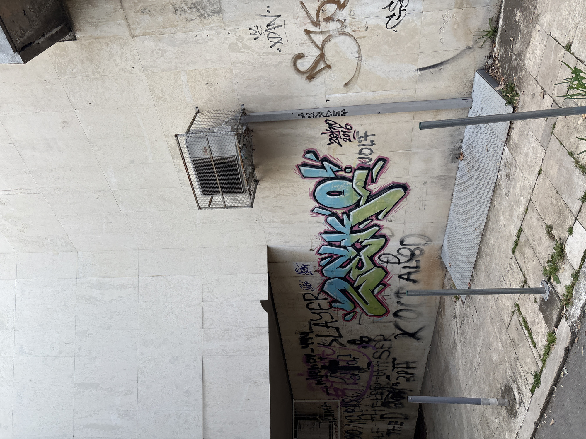 The image depicts an outdoor scene, likely a public walkway or alleyway, with a textured concrete wall on the left and a paved surface on the right. The wall is heavily adorned with graffiti in various styles and colors. Prominently featured is a large, colorful graffiti piece in blue, green, and pink, with sharp, angular letters. Below this, more graffiti in black and purple is visible, including the word "SLAYER" and what appears to be a date, "2011". Further up the wall, there are smaller tags and scribbles.
An outdoor air conditioning unit is mounted on the wall, enclosed in a metal cage. Two gray metal poles, possibly part of a railing or bike rack, are present in the foreground, extending horizontally across the frame. The paved area shows signs of wear and is interspersed with patches of grass and debris, suggesting an urban or semi-urban environment. The lighting suggests it is daytime, with no specific weather conditions discernible. The overall impression is one of urban decay and street art.