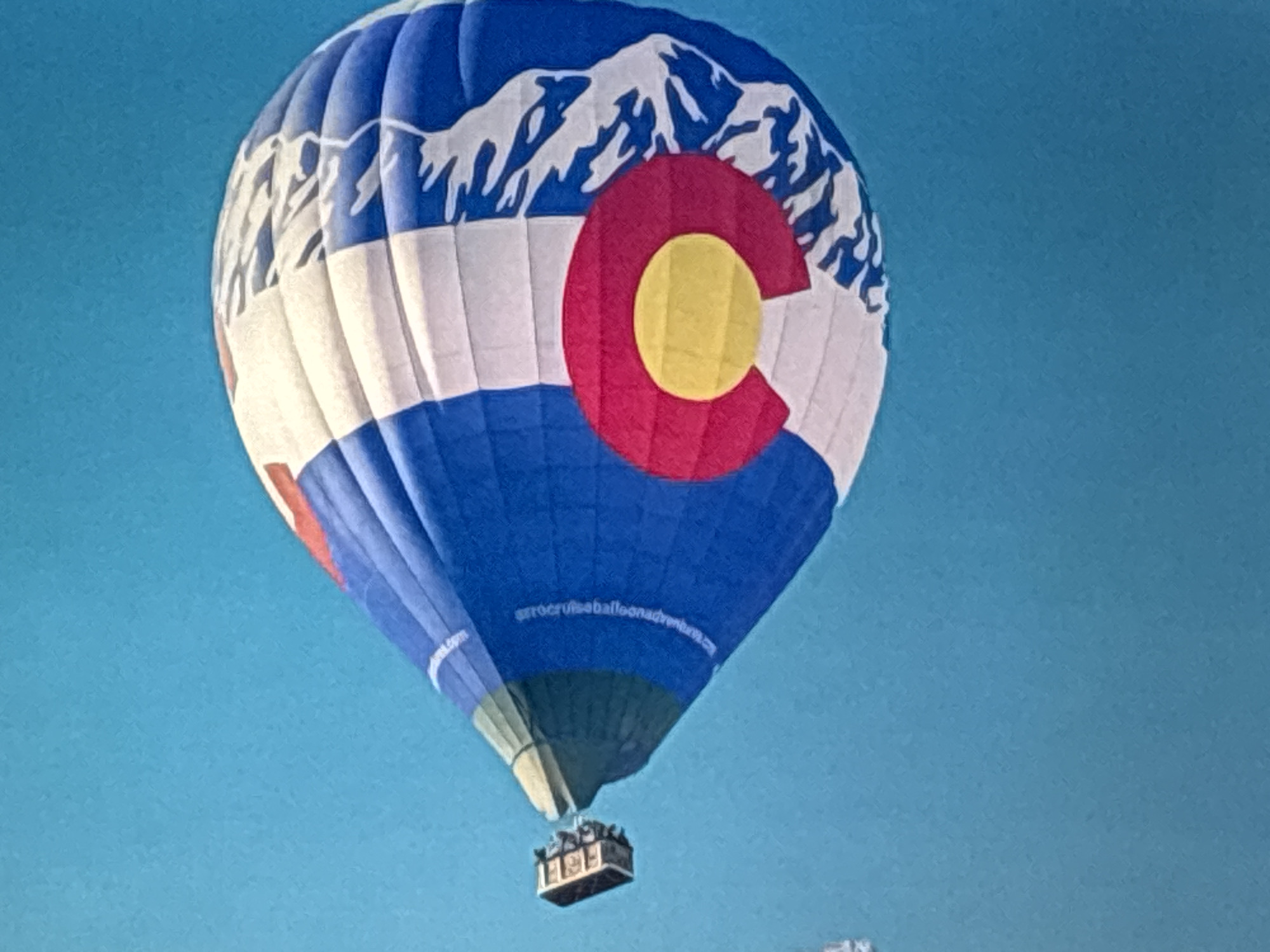A hot air balloon with a Colorado-themed design is captured in flight against a clear blue sky. The balloon's design features a stylized depiction of snow-capped mountains at the top, followed by horizontal stripes of white, blue, and a portion of red. Prominently displayed on the balloon is a large red "C" with a yellow circle inside, representing the Colorado state flag. Below the main body of the balloon, a basket is suspended, containing several figures who appear to be passengers. The basket is dark-colored, and the figures inside are small and indistinct due to the distance. The overall scene suggests a recreational hot air balloon ride.
There is visible text on the lower portion of the balloon, appearing to be a website address: "w.crocruiseballoonadventures.com". The bright, clear sky suggests it is daytime, likely with good weather conditions suitable for ballooning. The image focuses on the balloon, with the vast expanse of the sky serving as the backdrop. The composition emphasizes the balloon as the central subject, showcasing its unique design and its ascent into the atmosphere.