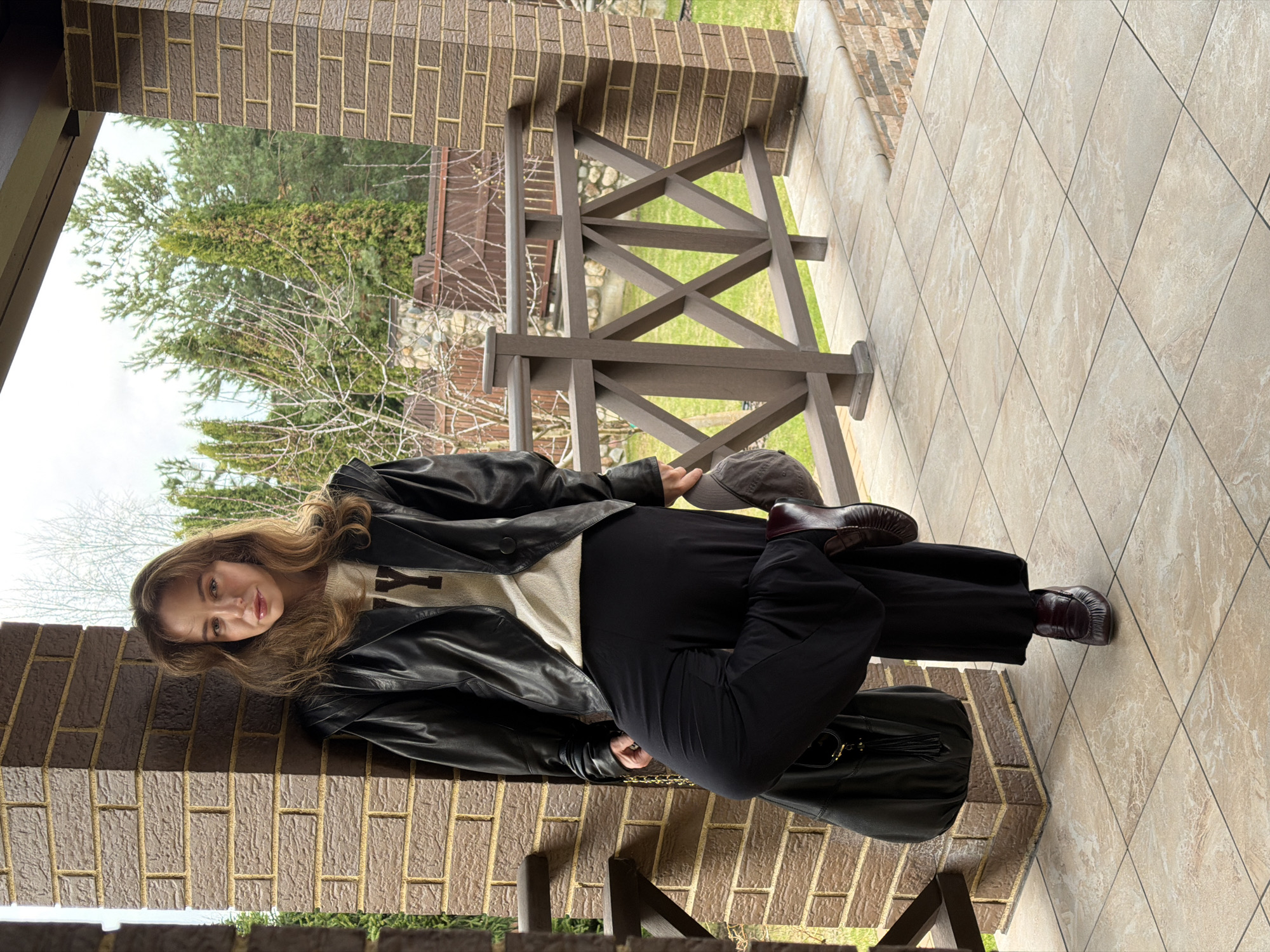 A young woman leans against a brick column, likely on a porch. She is wearing a black leather jacket over a light-colored sweater, black pants, and dark loafers. She holds a gray cap in her hand. A dark bag with gold chain detail sits on the ground near her. The background includes a wooden railing, a view of trees, and tiled flooring. The location is possibly Mishutkino, Russia, but this cannot be confirmed from the image.