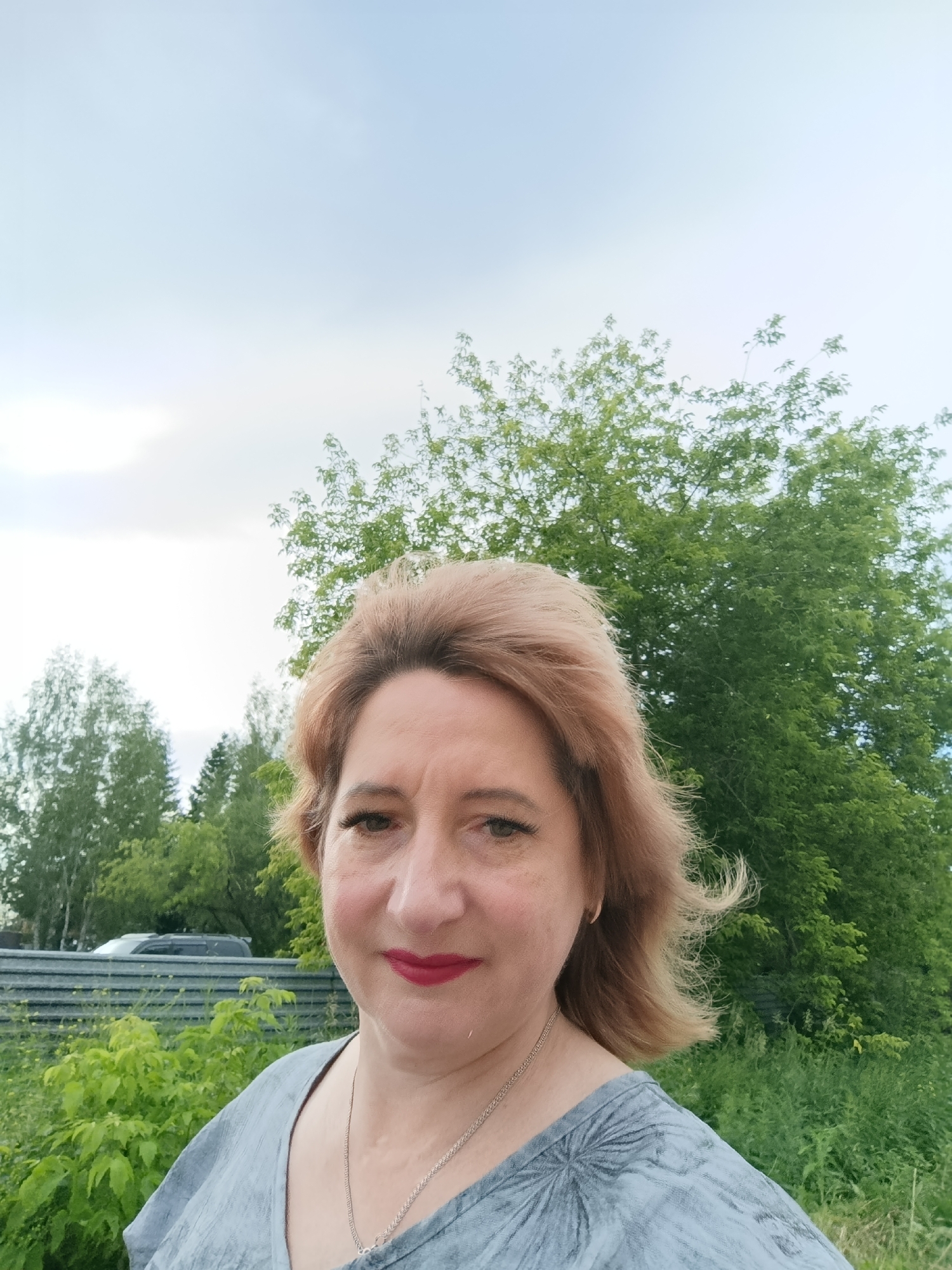 A woman with short, blonde hair and red lipstick takes a selfie outdoors. She is wearing a grey shirt and a silver necklace. Behind her, there are green trees and a grey fence. In the background, a car is visible. The sky is cloudy. The image is taken in Tugulym, Russia.