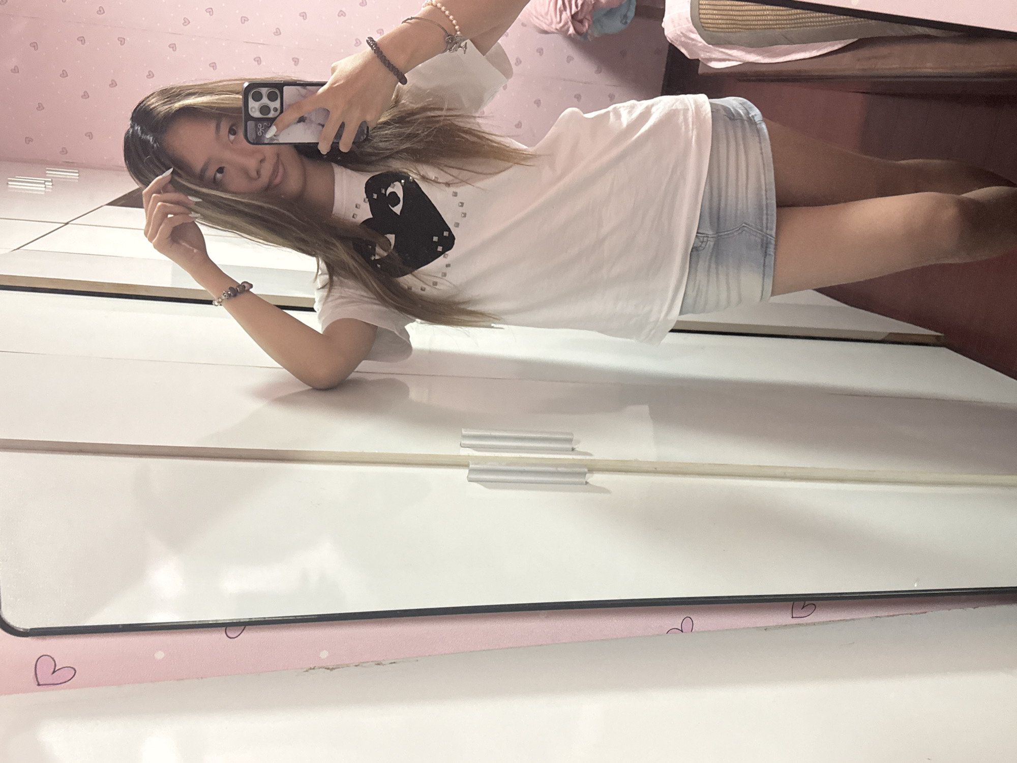 A young woman stands in front of a mirror, taking a selfie with her smartphone. She wears a white t-shirt with a black heart design and light-wash denim shorts. The background shows a pink wall with a repeating heart pattern and a partially visible bed with a woven surface. The mirror's frame has a pink edge with small heart designs. The photo was likely taken indoors. There is no indication the photo was taken in Shapingba District, China.