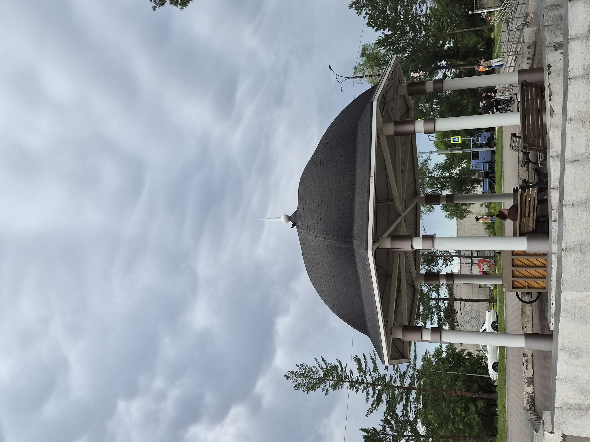 The image shows an outdoor gazebo with a dark, domed roof supported by white and brown columns. The gazebo is situated in what appears to be a park or public square, with benches inside. In the background, there are tall pine trees, a paved road, and several people. A white car is parked on the side of the road. Further in the background, a blue truck and a street light are visible. The sky is overcast with dramatic, grey clouds.
Specifically, the image depicts a gazebo in the city of Arseniev, Russia. The architecture and surrounding vegetation are consistent with urban public spaces in the Russian Far East. The presence of a blue light truck, typical of those used in Russia, further supports this location. The overall atmosphere suggests a slightly cool or damp day due to the cloudy skies.