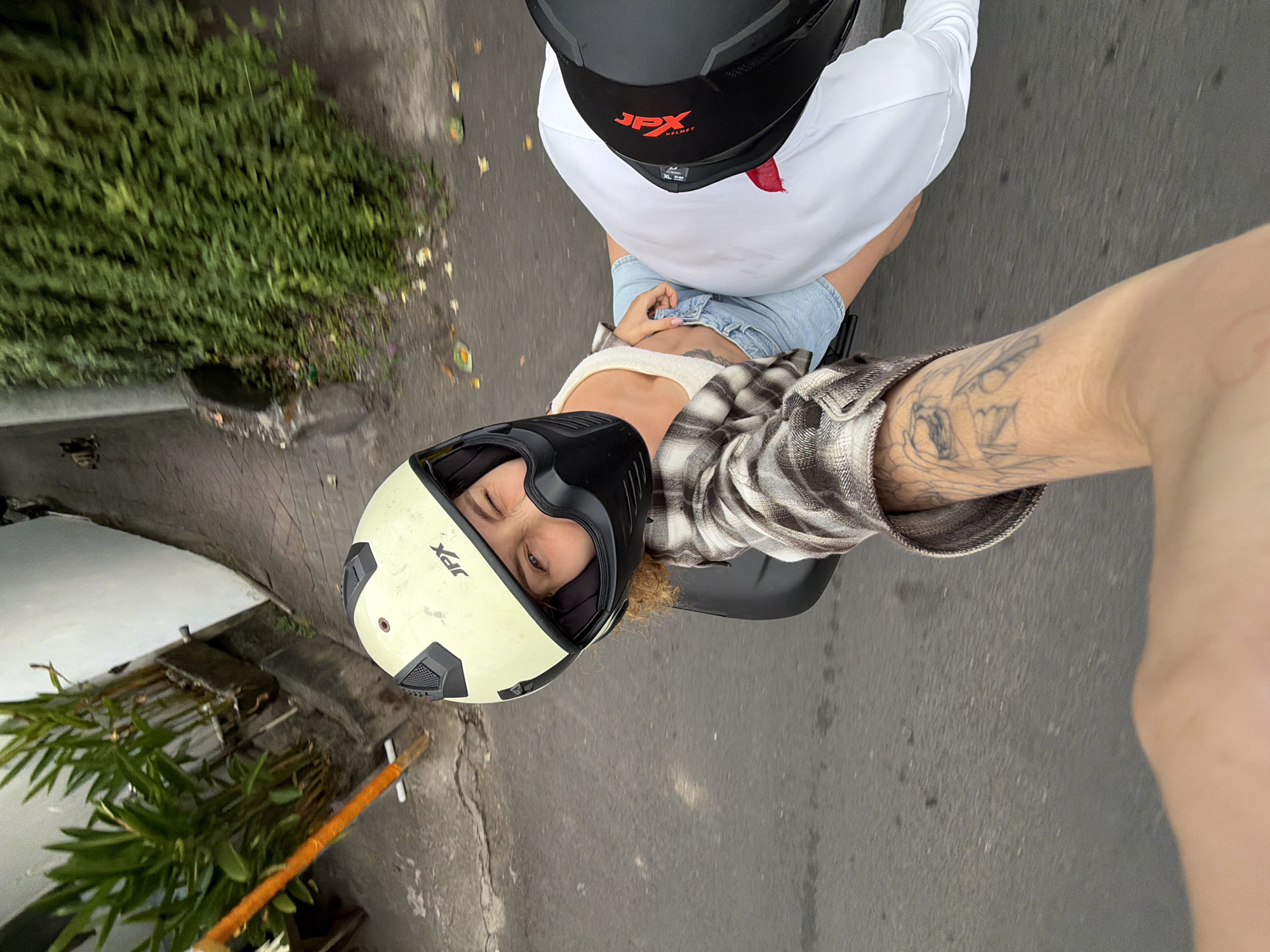 This is a first-person perspective, slightly over-the-shoulder shot of two people riding a scooter on a paved road, likely in North Kuta, Indonesia. The primary subject is the person in the foreground, wearing a cream-colored helmet with "JPX" visible in red and black text on the side. They are also wearing a plaid shirt over a white tank top and denim shorts. Their left eye is winking, and they are looking towards the camera. A tattoo is visible on their arm.
Behind them, the second person is wearing a black helmet, also with "JPX HELMET" visible in red. They have on a white t-shirt and are seated in front of the person taking the photo. The scooter seat is visible between them.
The scene includes a lush green bush on the left side of the road, and some fallen leaves are scattered on the asphalt. The lighting suggests it is daytime, possibly with overcast skies as there are no strong shadows. The overall impression is a casual, on-the-go moment, captured as a selfie while riding the scooter. The road surface appears somewhat worn.