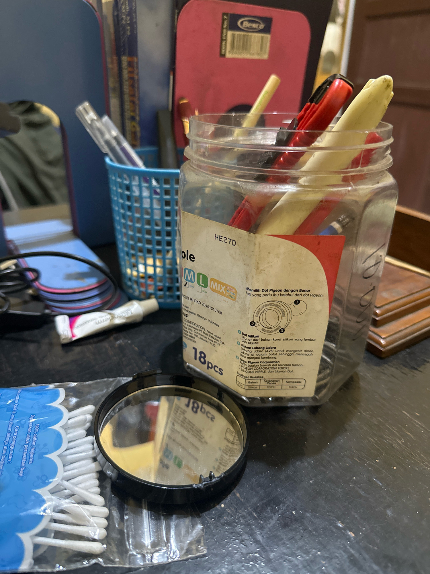 A close-up, eye-level shot shows a cluttered tabletop with various objects. In the foreground, a package of cotton swabs is partially visible on the left, alongside a small, round black mirror. Behind the mirror, a tube of white cream with pink and purple accents is lying on its side. Dominating the center of the image is a clear plastic jar filled with pens, markers, and other writing utensils. The jar has a label with text in Indonesian, including "HE27D," "Pigeon," and "18pcs." To the left of the jar, a blue plastic basket holds more pens. In the background, a pink object with the "Besto" logo is visible, along with other indistinct items and what appears to be a wooden surface. The lighting is dim, casting shadows on the tabletop, which has a textured, dark surface. The image is captured with a shallow depth of field, blurring the background elements. There is no discernible indication of the specific location within Saruni, Indonesia, or any people or actions.