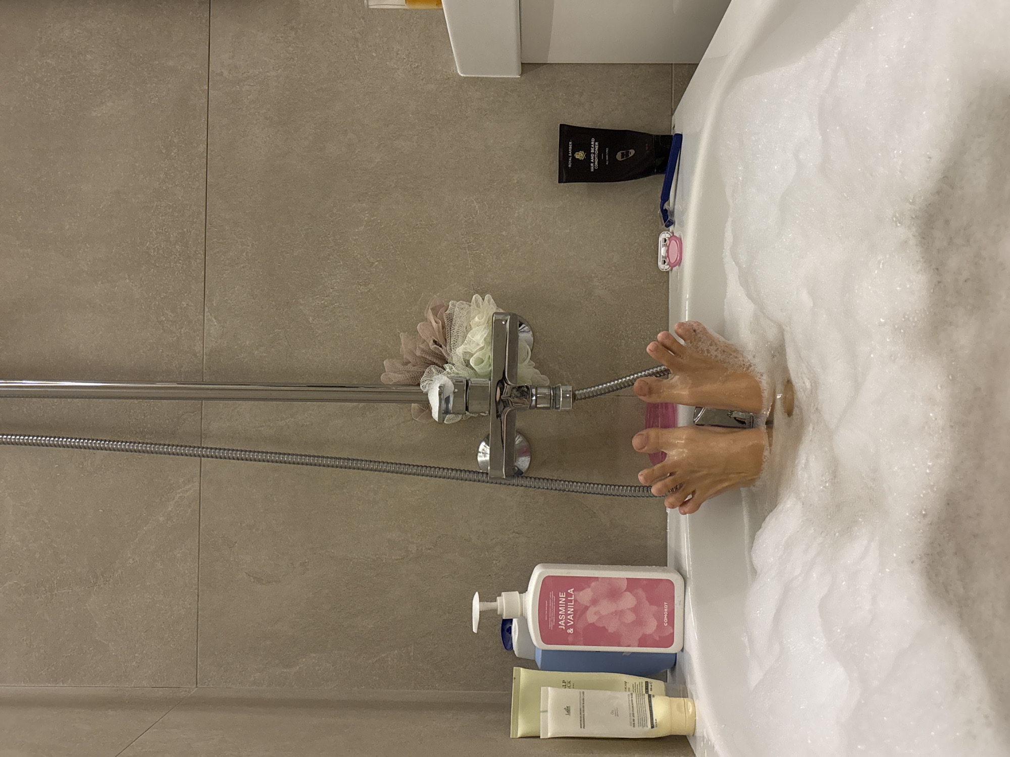 The image shows an overhead view of a bathtub filled with bubbly bathwater. A person's feet are visible, submerged in the water. Several bottles of bath products, including "Jasmine & Vanilla" bubble bath by a brand called "Самокат", are lined up along the edge of the tub next to a shower head. A loofah is also visible. A black tube of "Royal Barber Hair and Beard Conditioner" and a pink razor also sit on the edge of the tub. The walls are tiled in a light brown color. The brand of bubble bath, "Самокат," is a Russian brand based out of Krasnoyarsk, Russia.