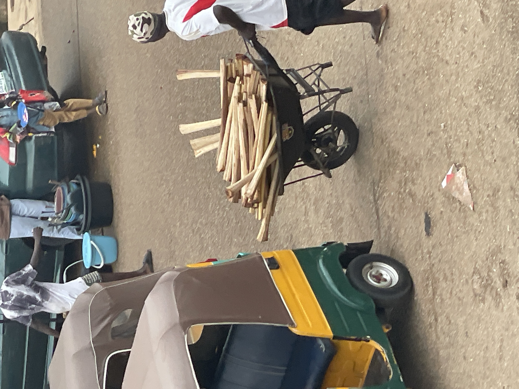 In an outdoor urban environment with a focus on a paved surface, a person is captured from the waist down, pushing a black wheelbarrow filled with a large quantity of cut wooden branches. The wheelbarrow has two wheels and a metal frame. To the left of the wheelbarrow, a green and yellow auto-rickshaw is parked. The auto-rickshaw has a brown canopy over its passenger compartment. The background is blurred, but suggests vehicles and other people, implying a street or market setting. A person in a white shirt with red accents and a camouflage patterned hat is partially visible on the right side of the frame, walking away from the camera. Another individual, wearing a patterned shirt and shorts, is seen on the far left, partially obscured by the auto-rickshaw. The ground appears to be damp in some areas.