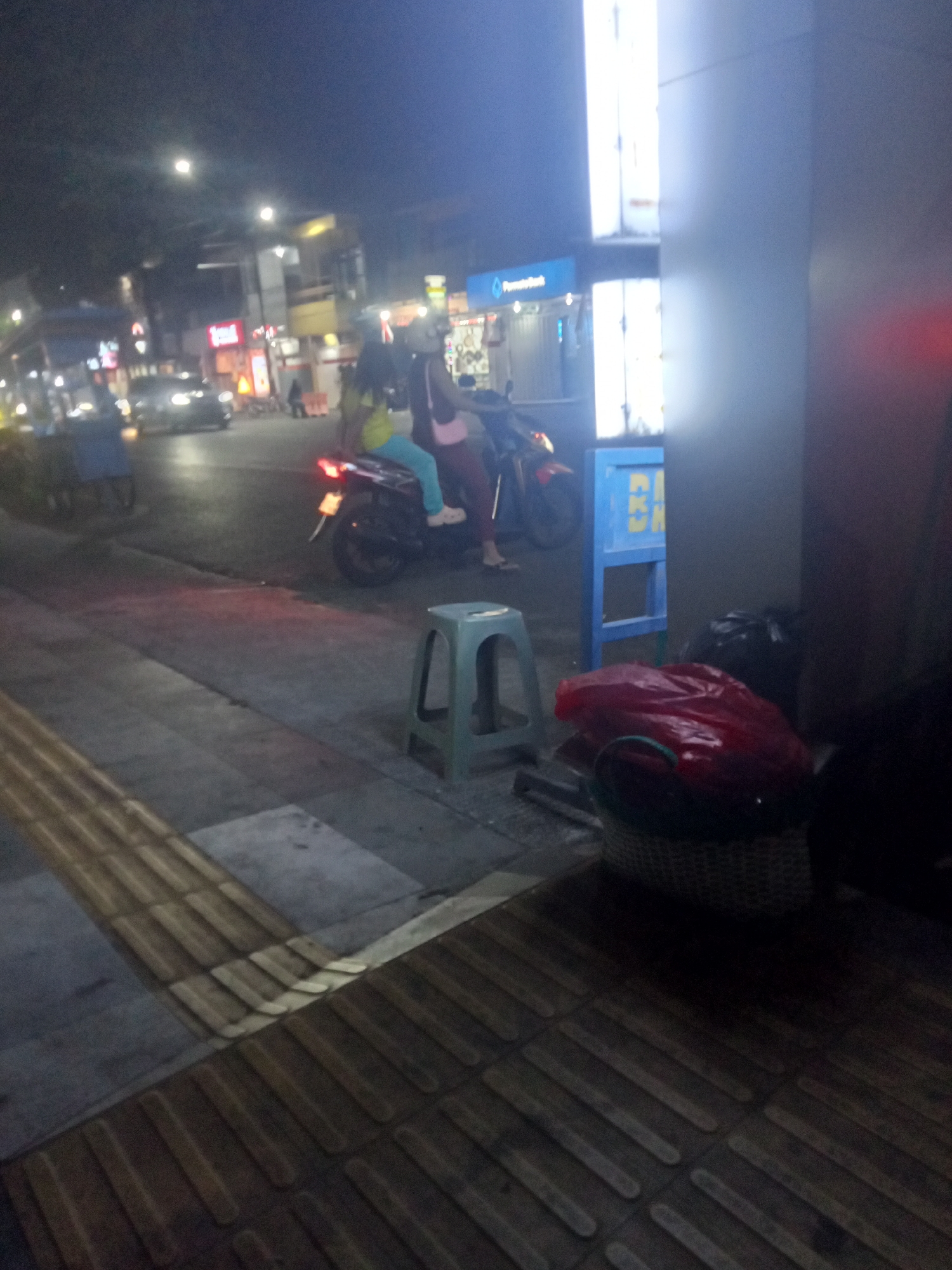 The image depicts a street scene at night in Probolinggo, Indonesia. In the foreground, a textured sidewalk with tactile paving is visible. To the right, a woven basket is filled with a red plastic bag and other items, next to a black trash bag. A small, light blue plastic stool is positioned on the sidewalk. Further back, a motorcycle with two occupants, a driver and a passenger, is parked or stopped on the road. The passenger, wearing a yellow top and blue pants, is partially visible. The motorcycle's brake light is illuminated. A blue stand with yellow lettering is situated behind the motorcycle. In the background, a building with illuminated windows and a sign for "Permata Bank" is visible. Other buildings and streetlights line the road, and a car with illuminated headlights is driving on the street. The overall scene suggests a typical urban environment at night, with activity on the street and in the adjacent buildings.