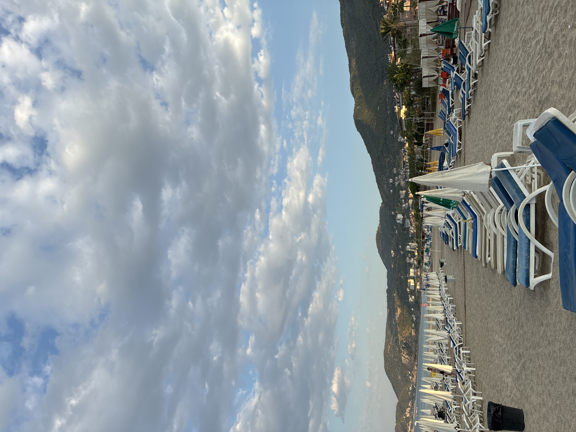 The image depicts a view of a beach in Alanya, Turkey, with rows of empty lounge chairs and umbrellas set up on the sand. The beach is situated in front of a coastline with lush green hills and mountains in the background. Buildings and palm trees are visible along the coast. The sky is partly cloudy with a mix of blue sky and white clouds. The scene appears calm, possibly during the early morning or late afternoon.